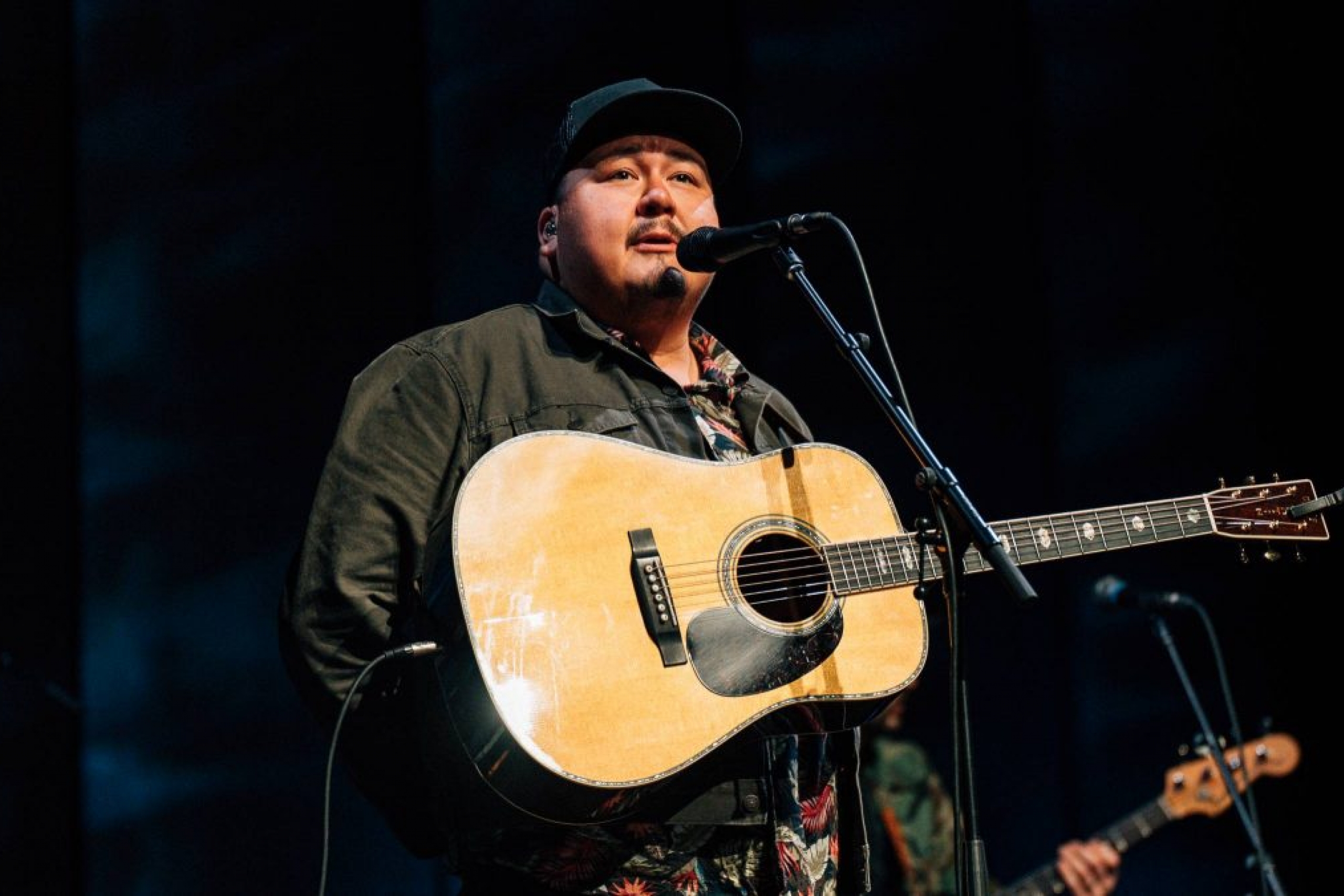 A man in a black baseball cap with a guitar strapped across his chest stands speaking into a microphone