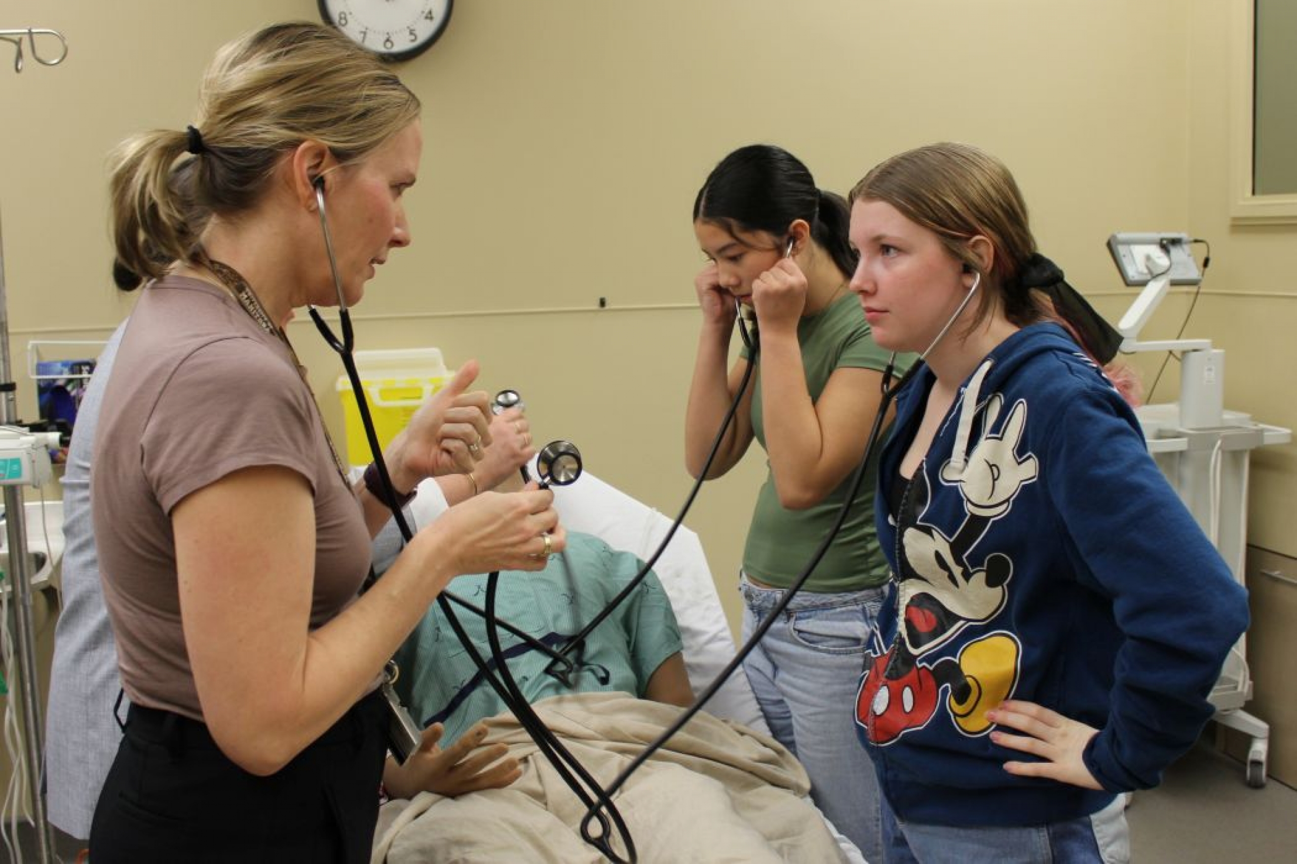 An instructor speaks to a student. Both are wearing stethoscopes. In the background is a student wearing a stethoscope and there is a training manikin on a hospital bed.
