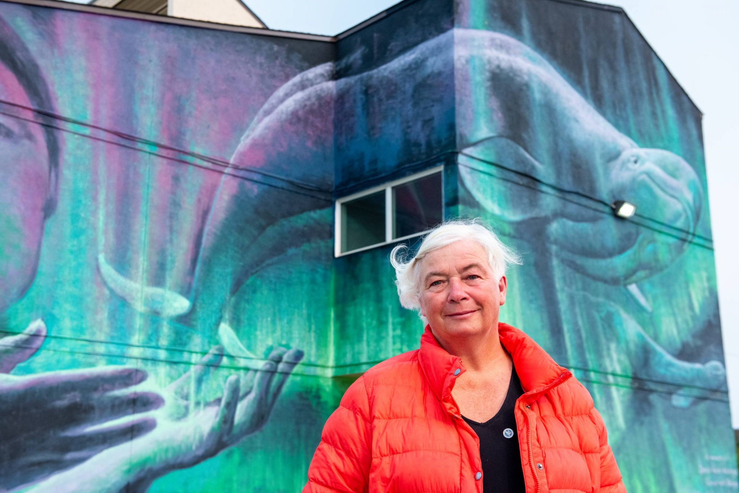 A white-haired woman in an orange coat stands in front of the outside of a building that has a mural on it