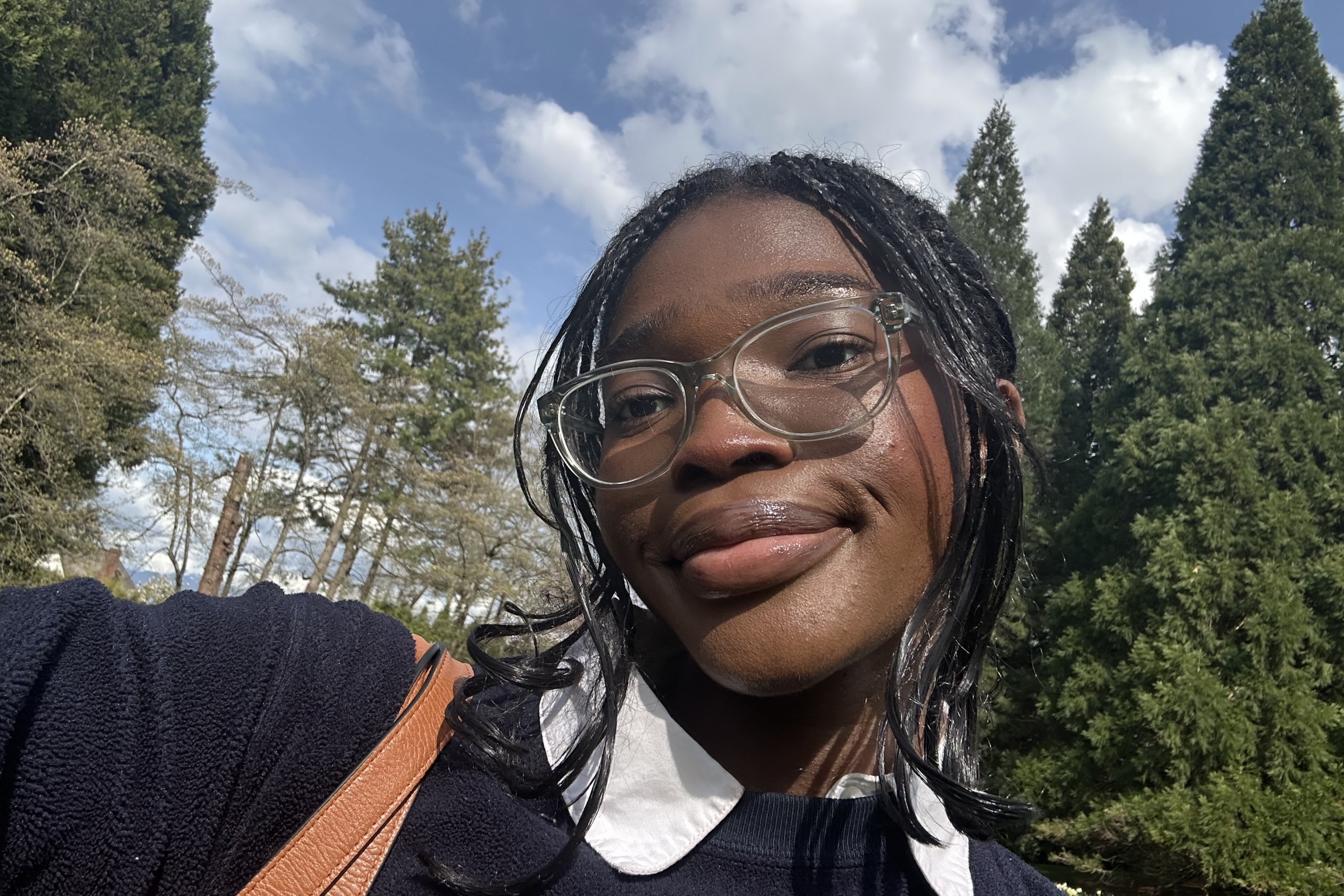 Elinam taking a selfie outdoors with pine trees in the background.