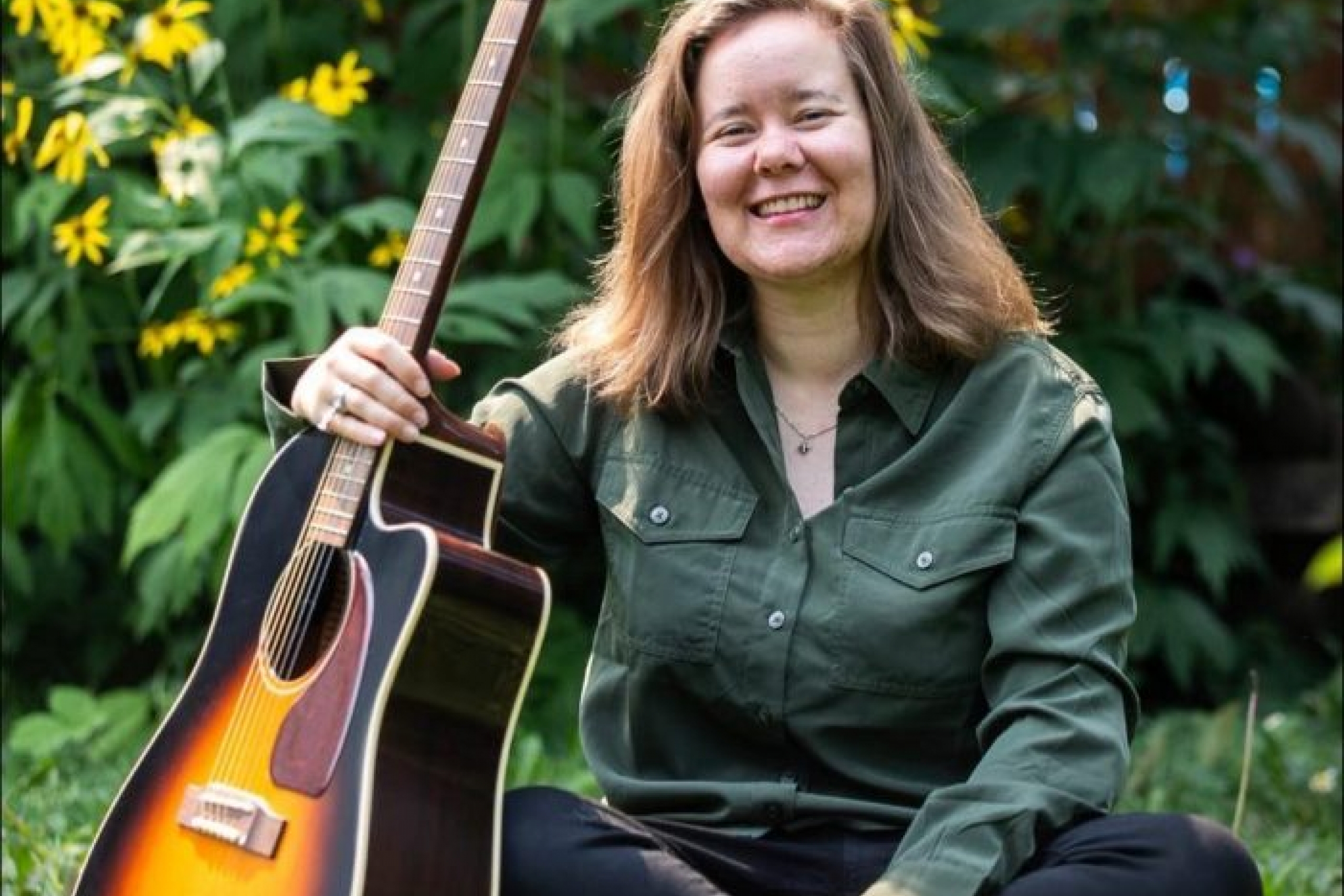 A smiling woman sits in grass with a guitar leaning against her legs.