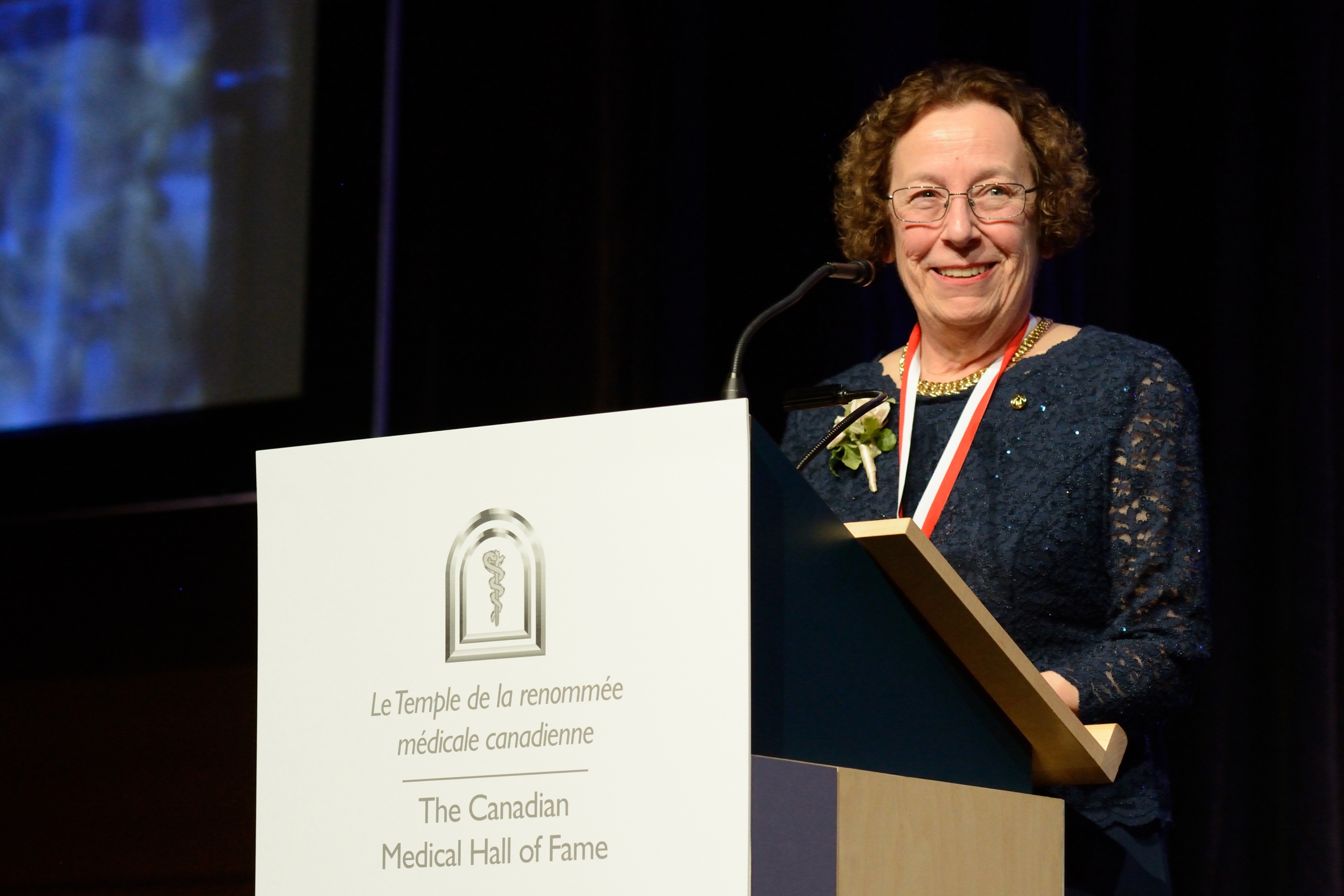Dr. Estelle Simons at a lectern. A sign on the lectern reads in French and English, The Canadian Medical Hall of Fame.
