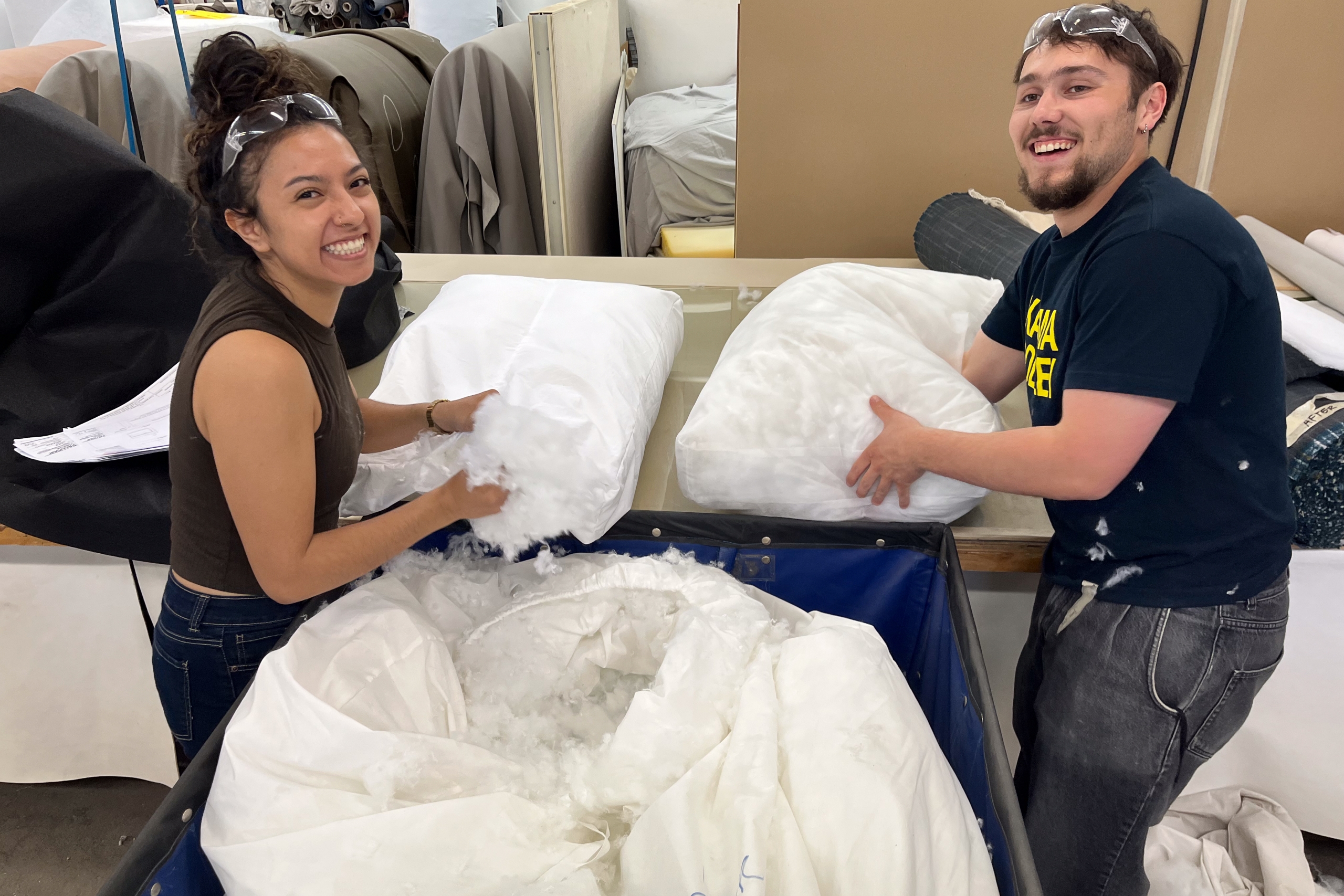 Two people smiling while stuffing pillows with filling in a workshop. They wear protective eyewear, surrounded by fabric and sewing equipment.
