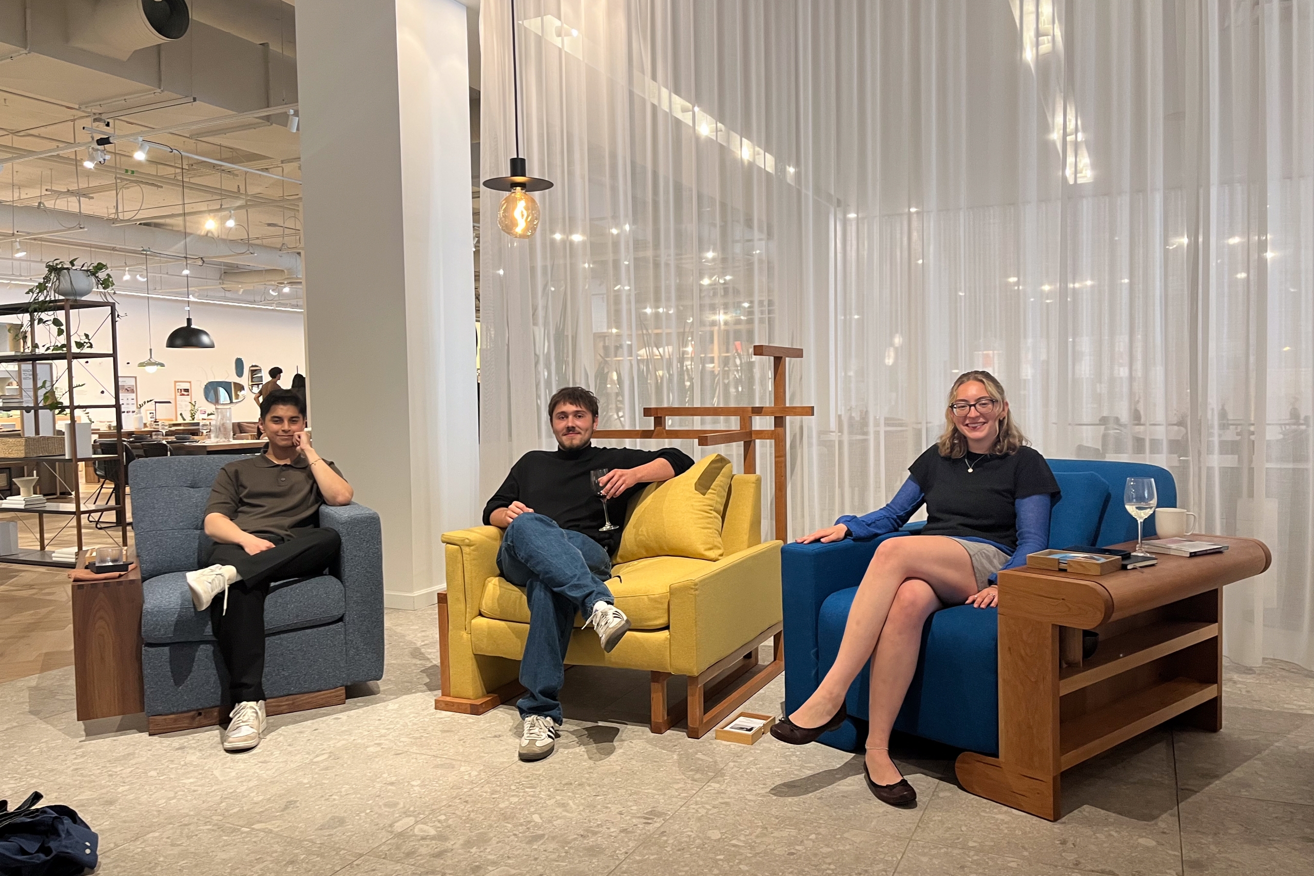 Three people sit on colorful armchairs—blue, yellow, and blue—in a modern, warmly lit room with sheer curtains; they appear relaxed and cheerful.