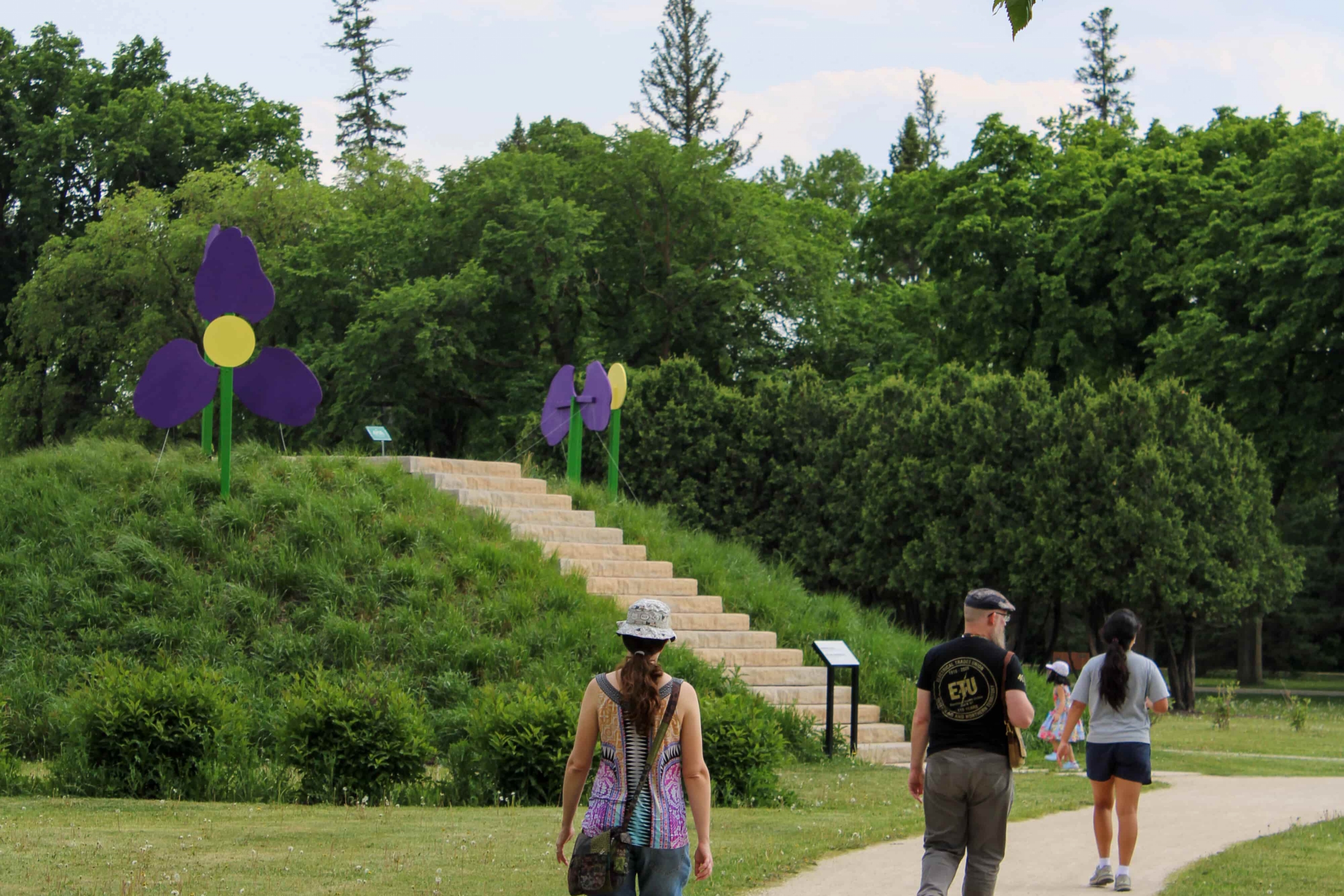 A scenic park path with people walking towards large purple and yellow flower sculptures on a grassy hill. Lush greenery and trees surround the area.