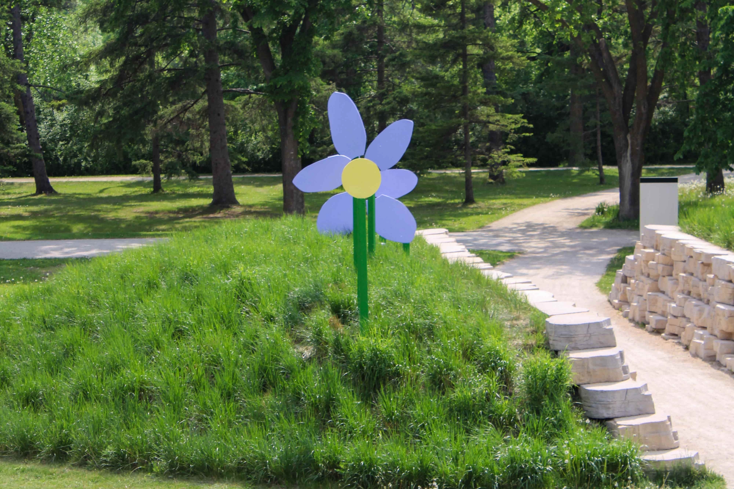 A large, colorful flower sculpture with purple petals and a yellow center stands on a grassy mound. It is surrounded by lush green trees and a paved pathway, creating a joyful and serene park scene.
