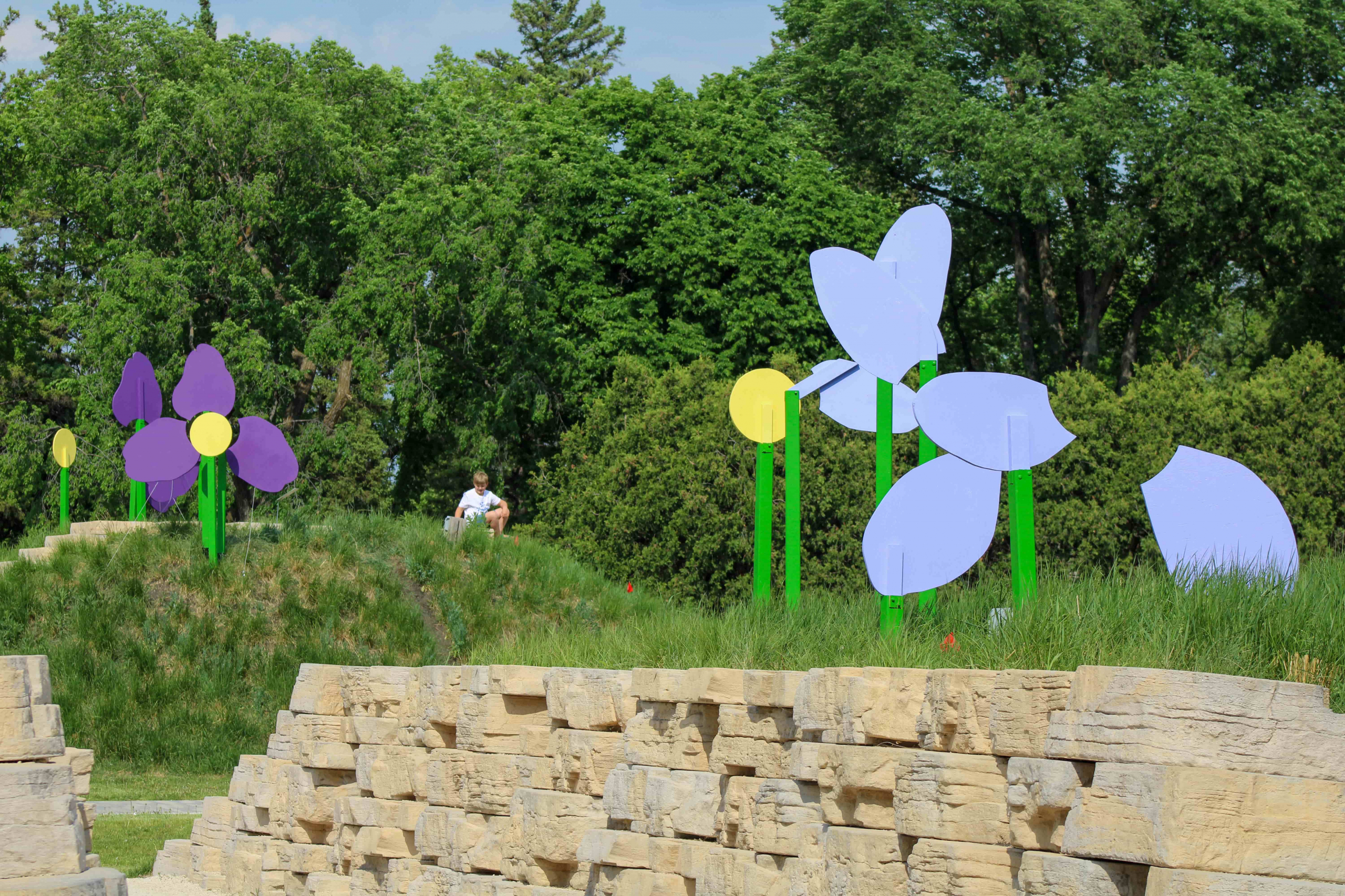 A grassy hill with colorful, oversized metal flowers, purple and yellow, against lush green trees. A person sits on the hill, creating a peaceful scene.