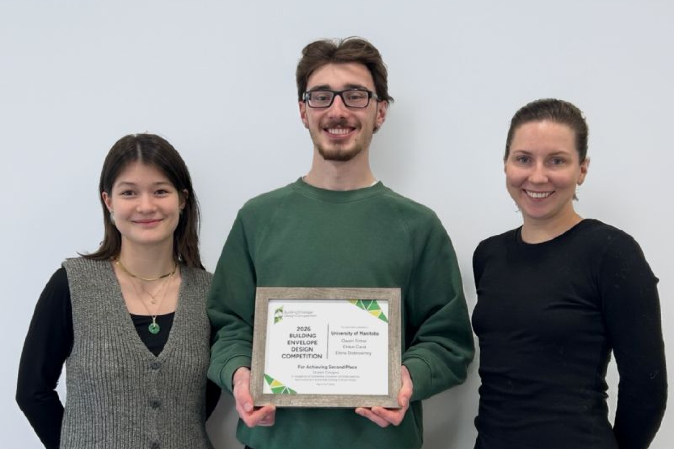 Three people stand against a plain white background, smiling. The person in the center holds a framed certificate, suggesting an award achievement.
