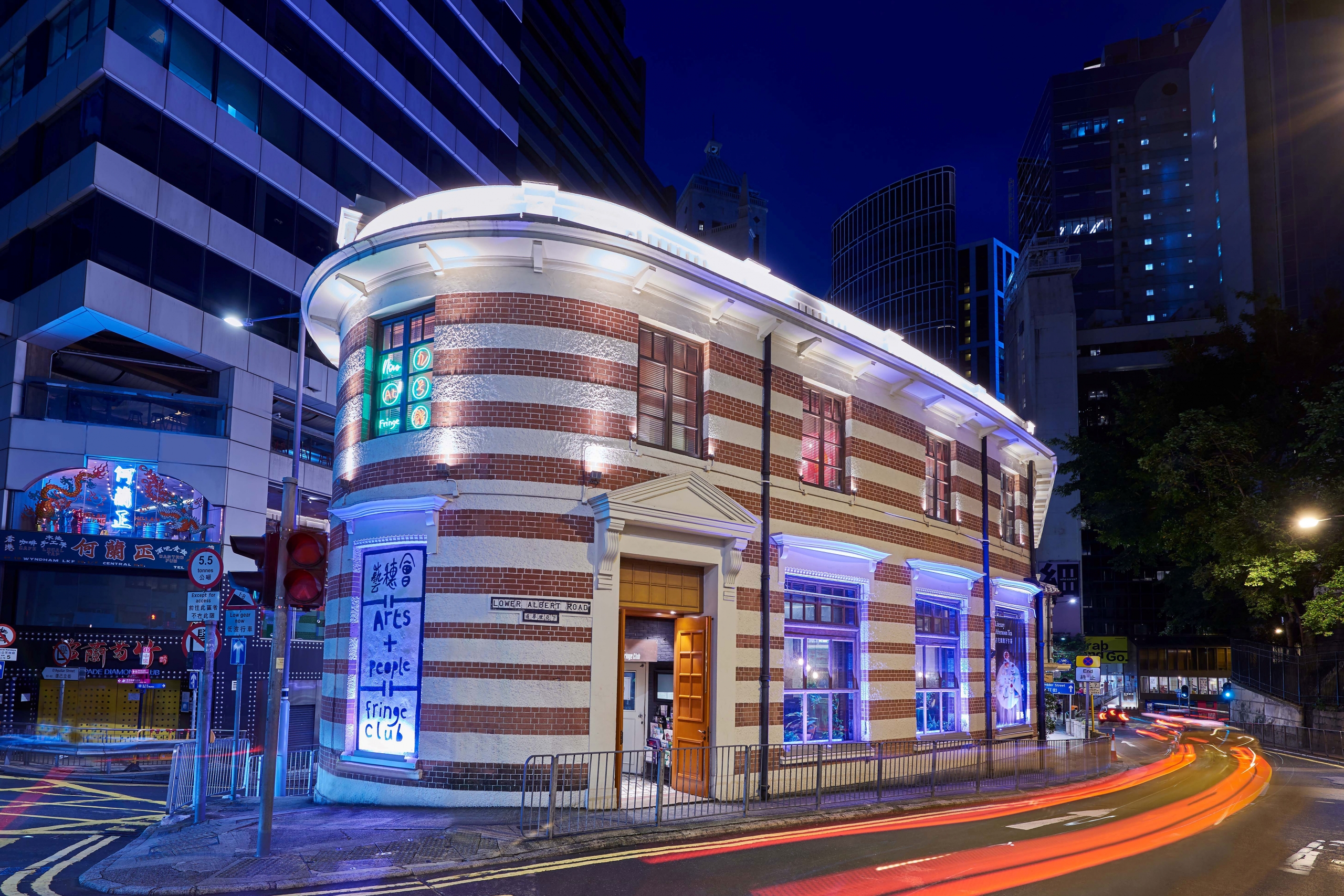 A lit, historical brick building with white trim on a city street at night, surrounded by modern skyscrapers. Light trails from passing cars add dynamic motion.