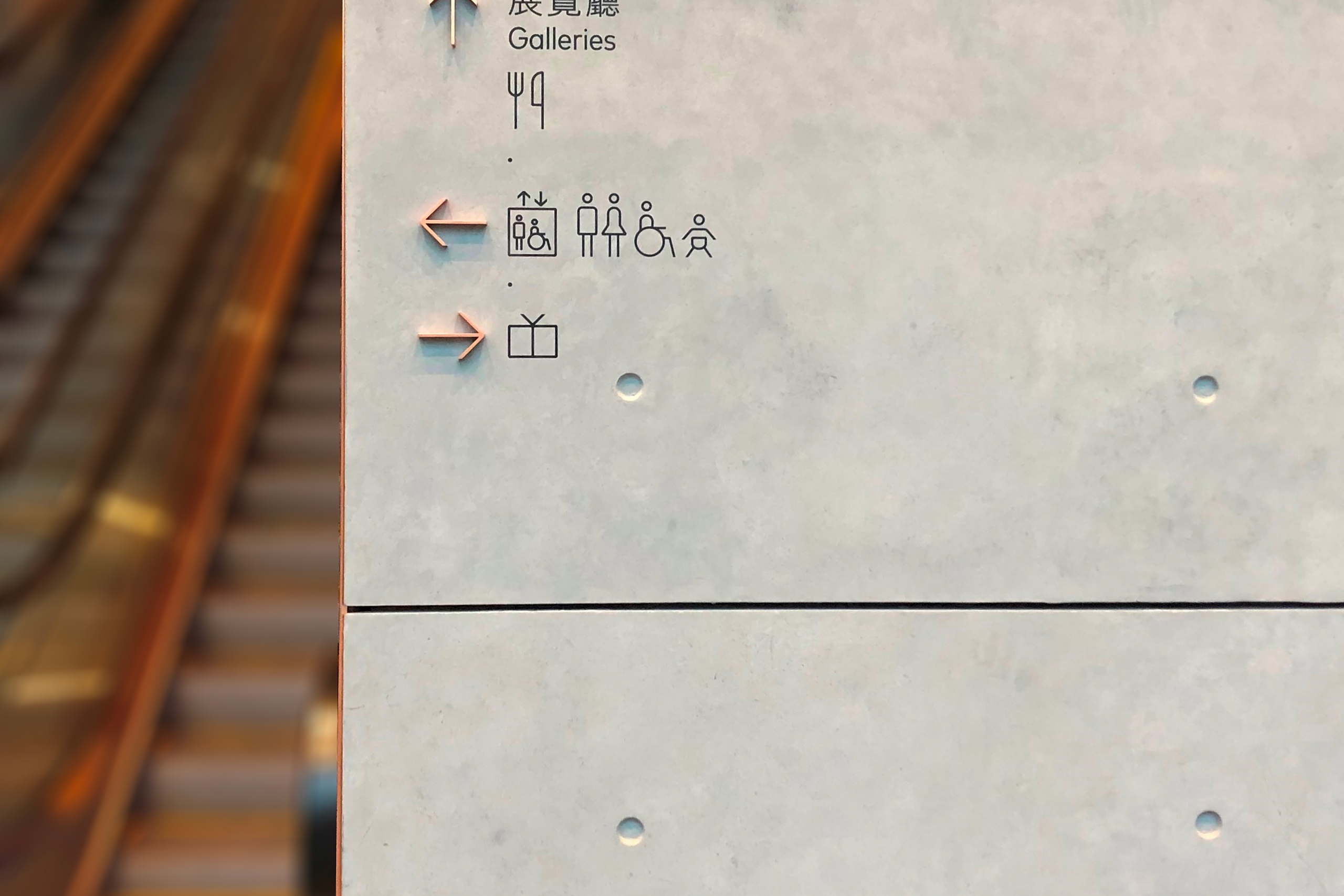 A concrete wall features directional arrows and icons for galleries, restrooms, and dining. An escalator is blurred in the background, enhancing depth.