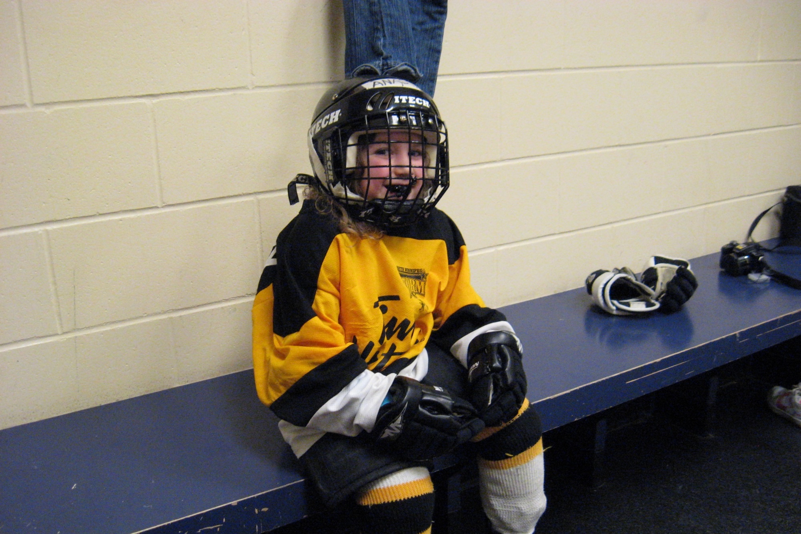 A young girl dressed in her hockey gear sits on the bench in the locker room.