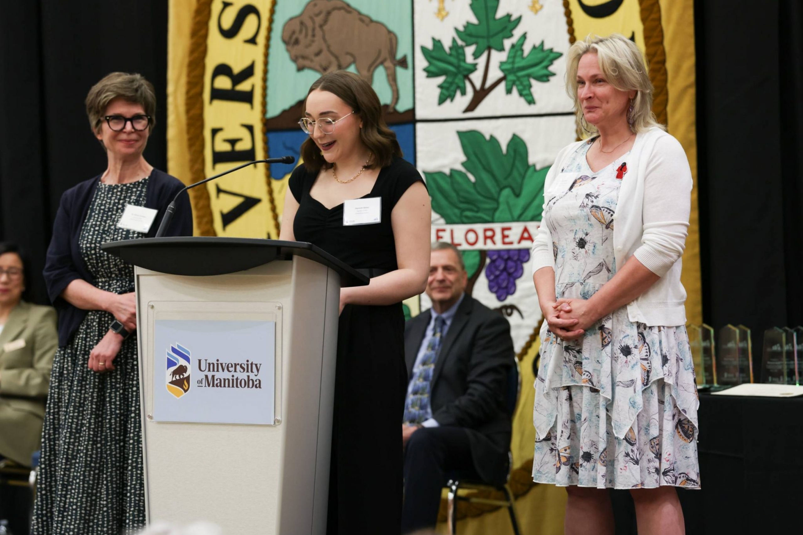 Two former educators are watching over a graduating student as she gives her speech.