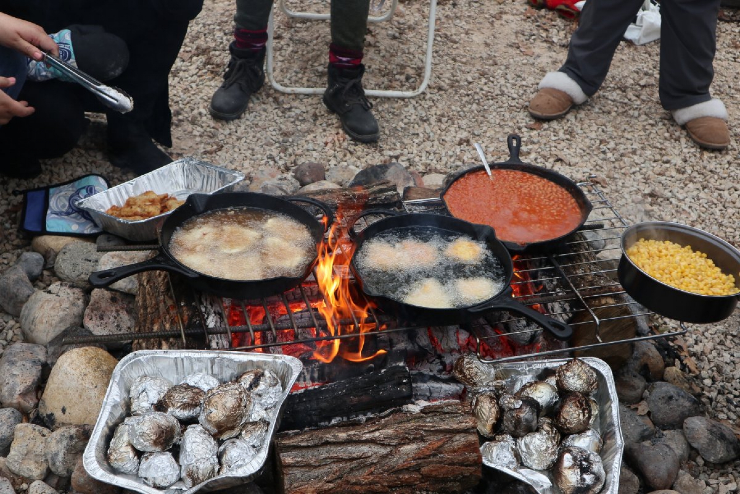 skillets over a fire, cooking fish, beans and corn.