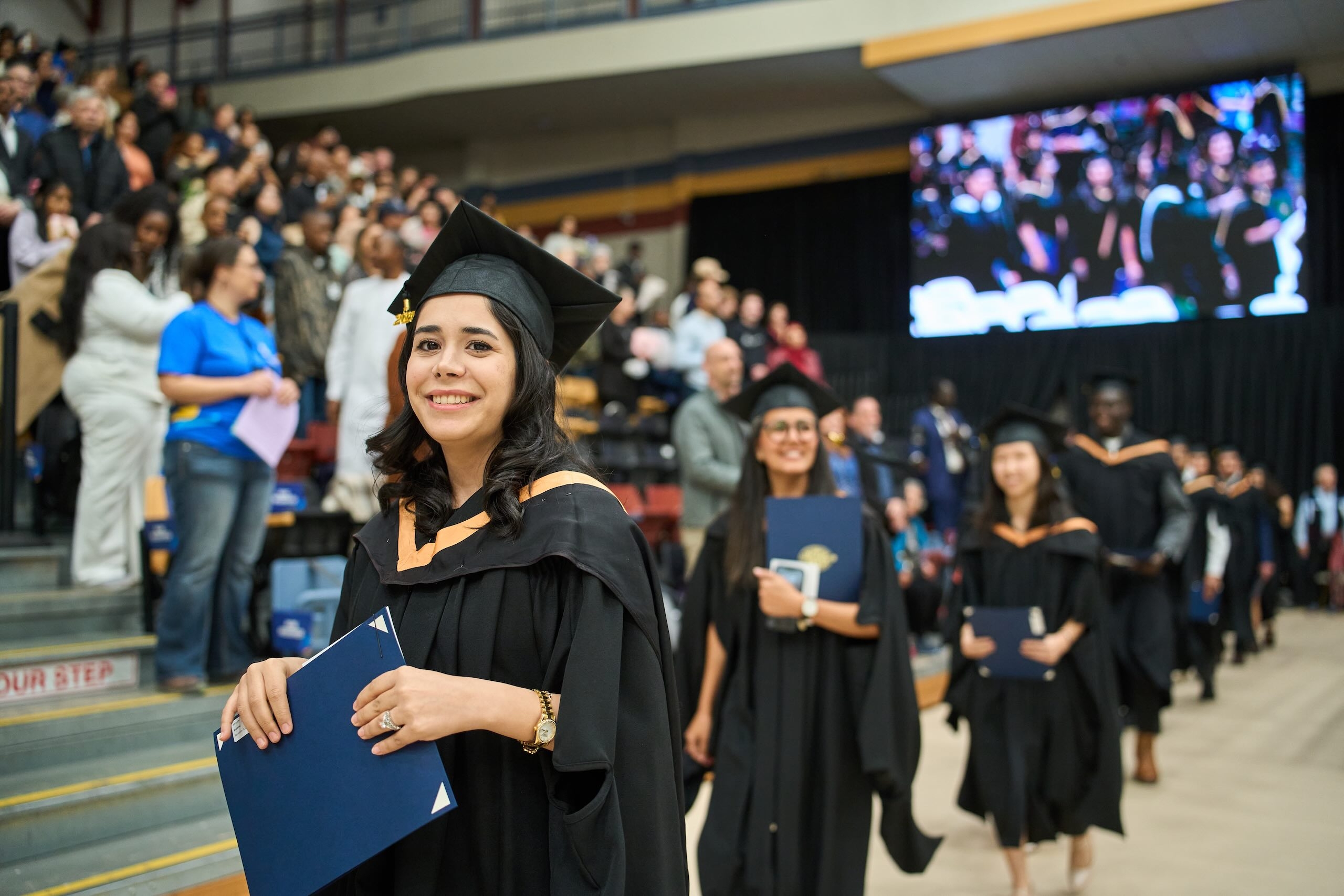 Young smiling woman in black grad cap and gown, holding diploma.