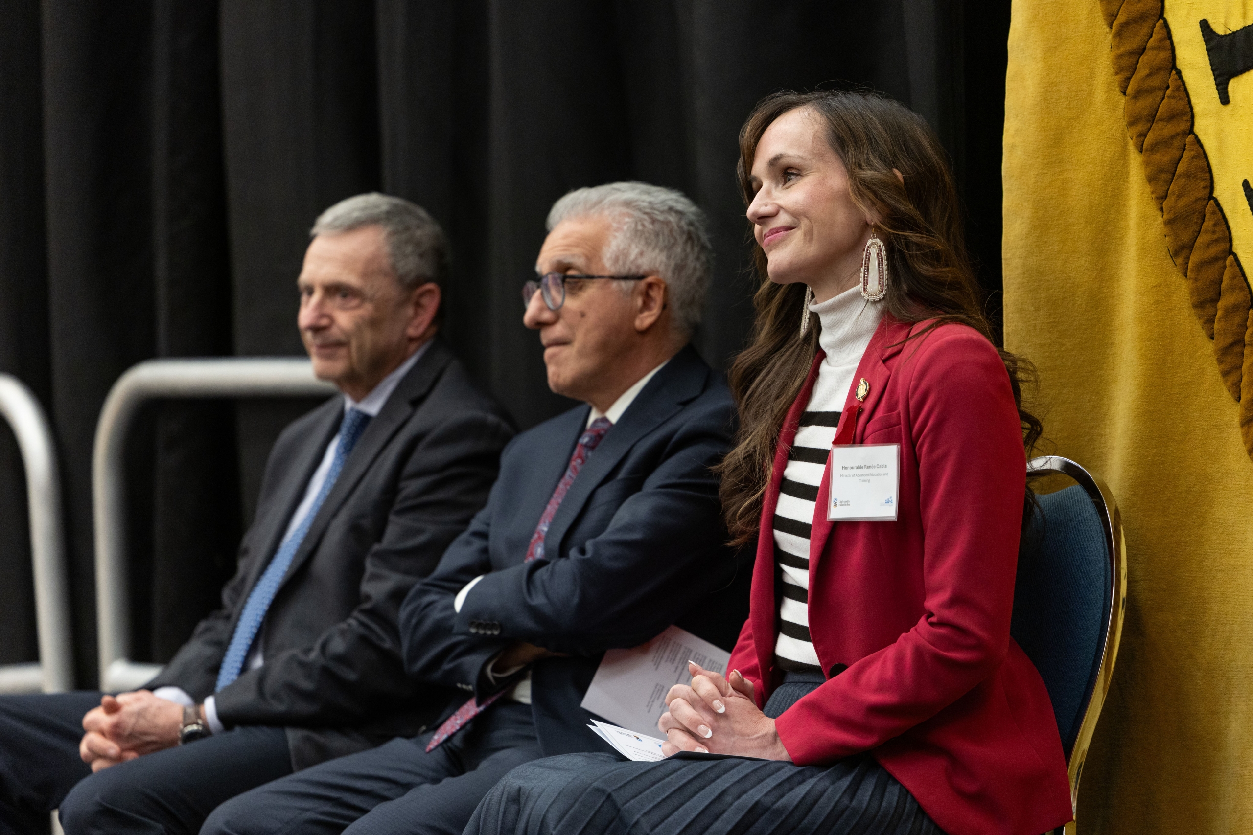 Dr. Mark G. Torchia, Dr. Michael Benarroch, and Honourable Renée Cable sit in formal attire at the back of the stage.