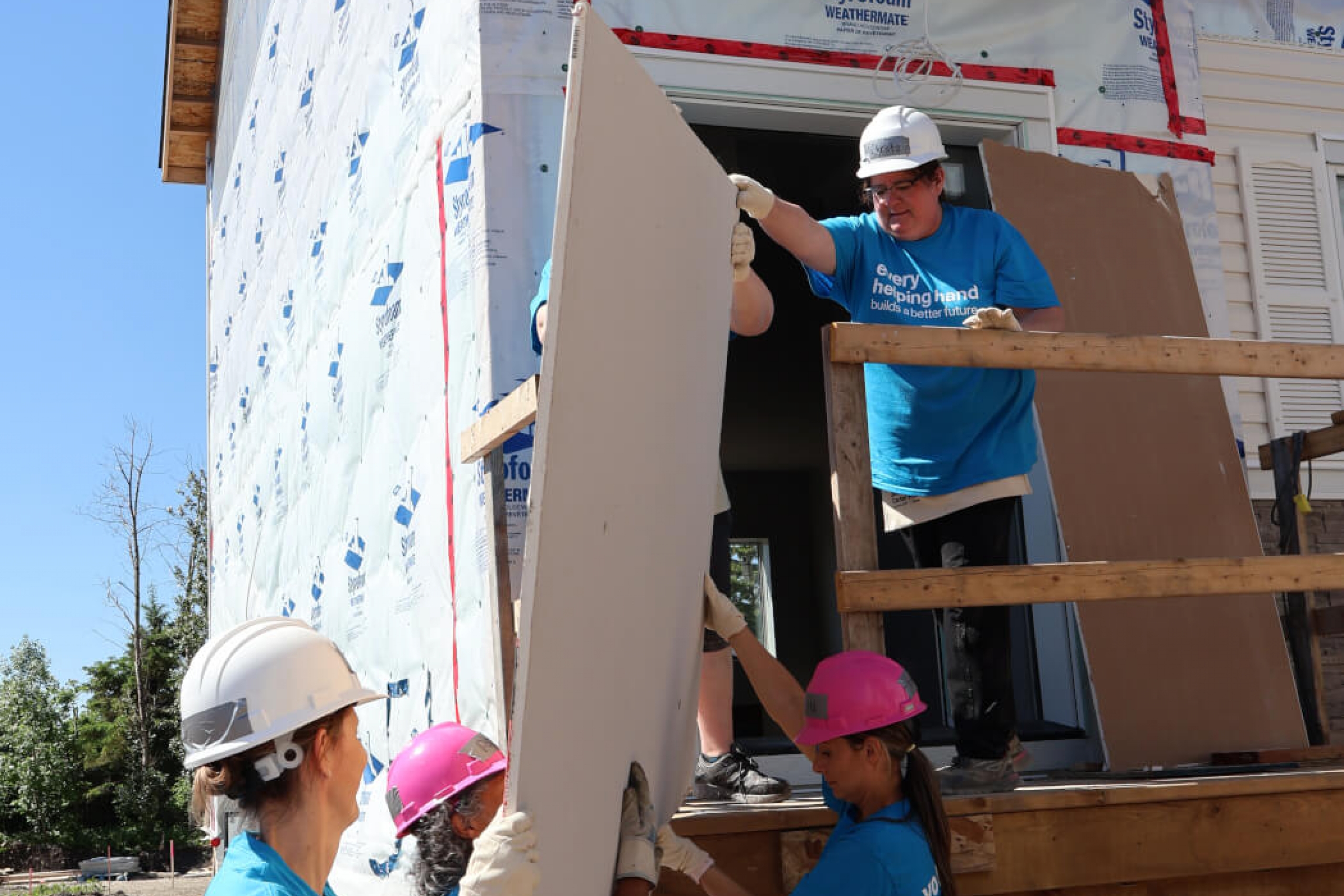 Three people on the ground pass a sheet of construction material to two people standing at the top of a staircase outside. They are all wearing hardhats.
