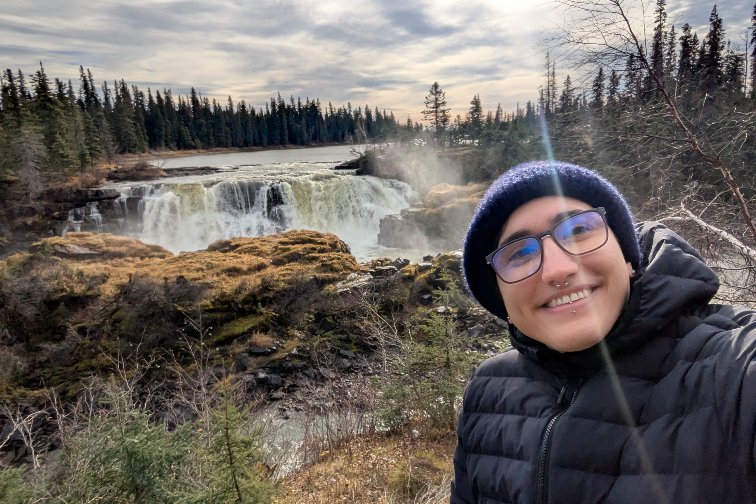 A person takes a selfie and smiles at the camera, in the background is Pisew Falls surrounded by pine trees.