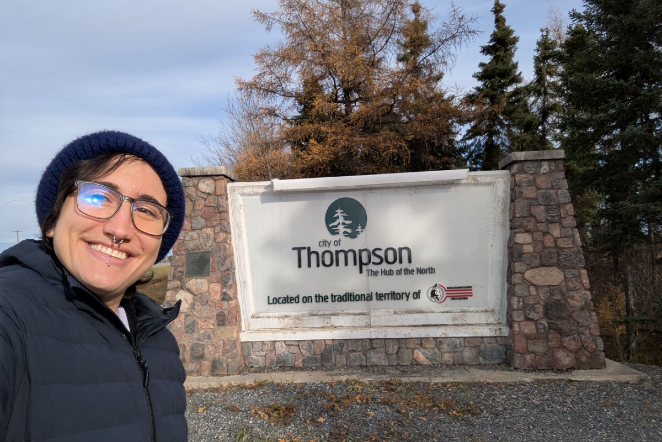 A person takes a selfie and smiles at the camera, in the background is a City of Thompson welcome sign.