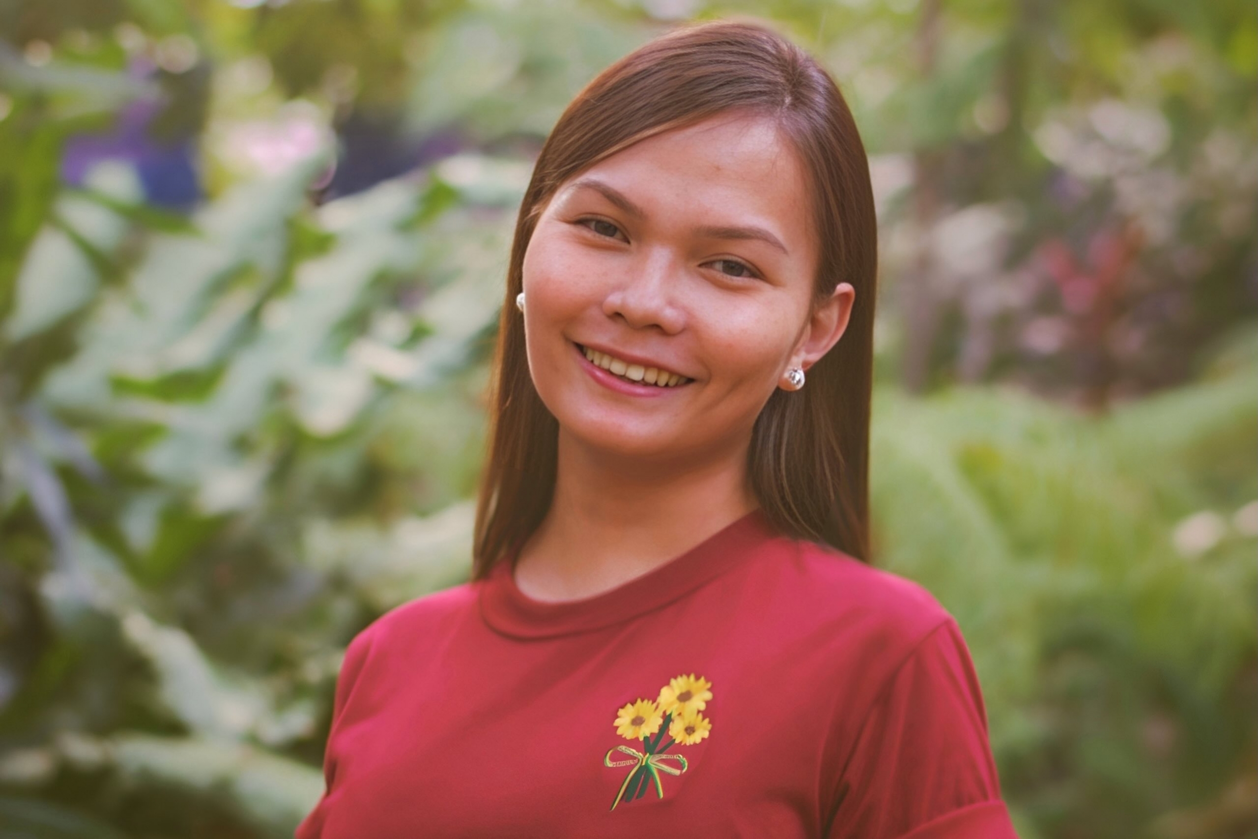 Young smiling woman with long hair and red shirt in front of greenery.