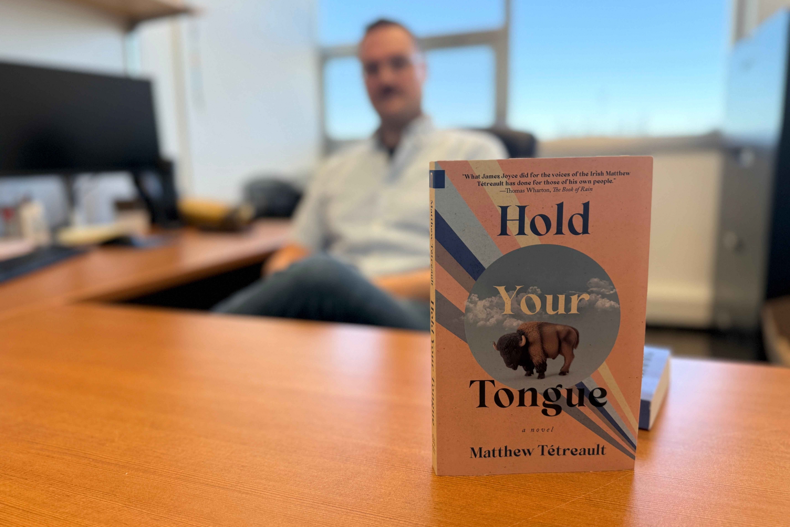 Novel stands upright on desk in focus, with male seated in blurred background.