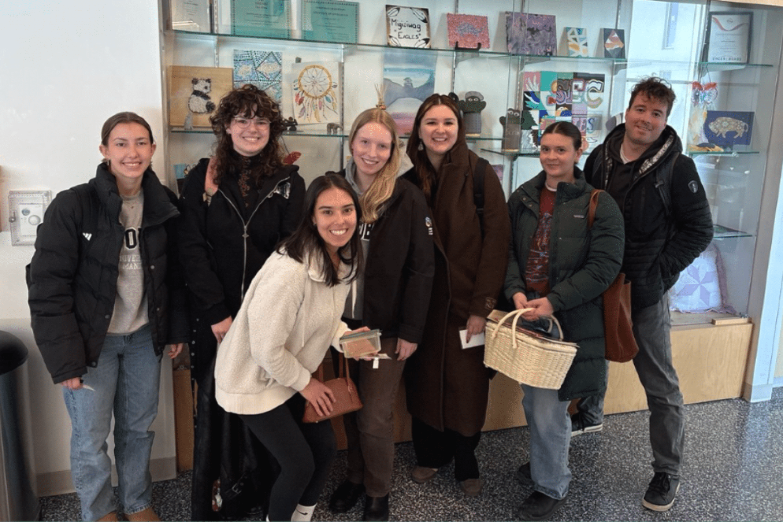 A group of students pose together in front of a display case of artwork in a campus building.