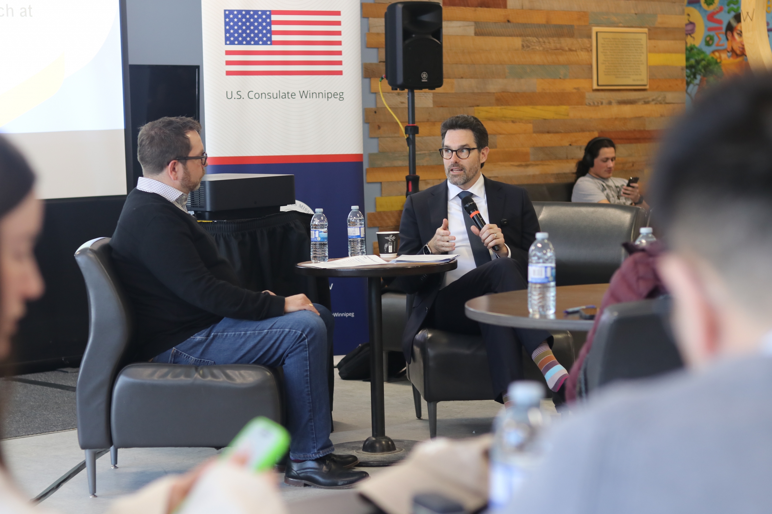Two speakers engaged in a fireside chat, seated in chairs before an audience.