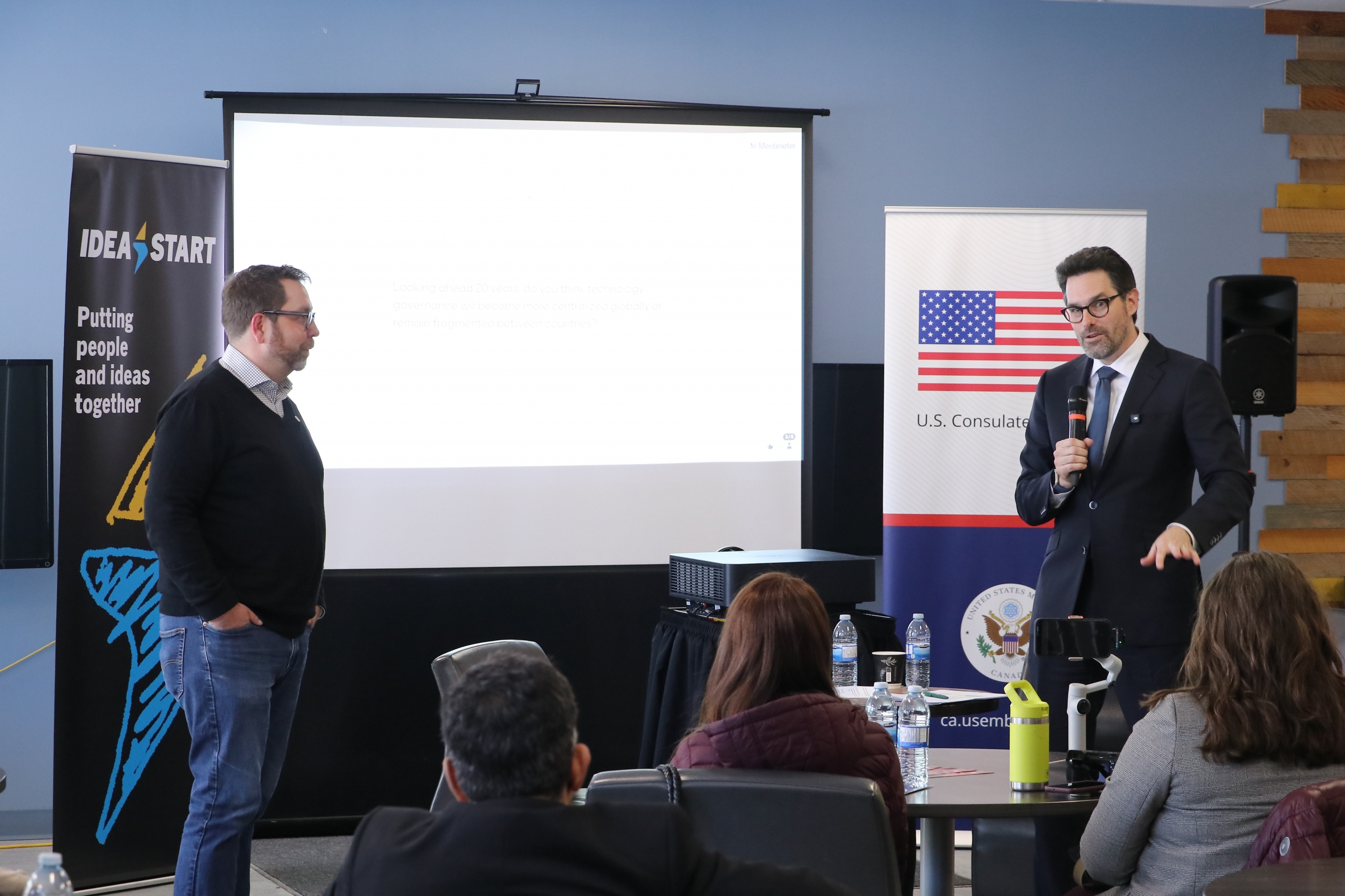 Two speakers engaged in a fireside chat, standing before an audience.
