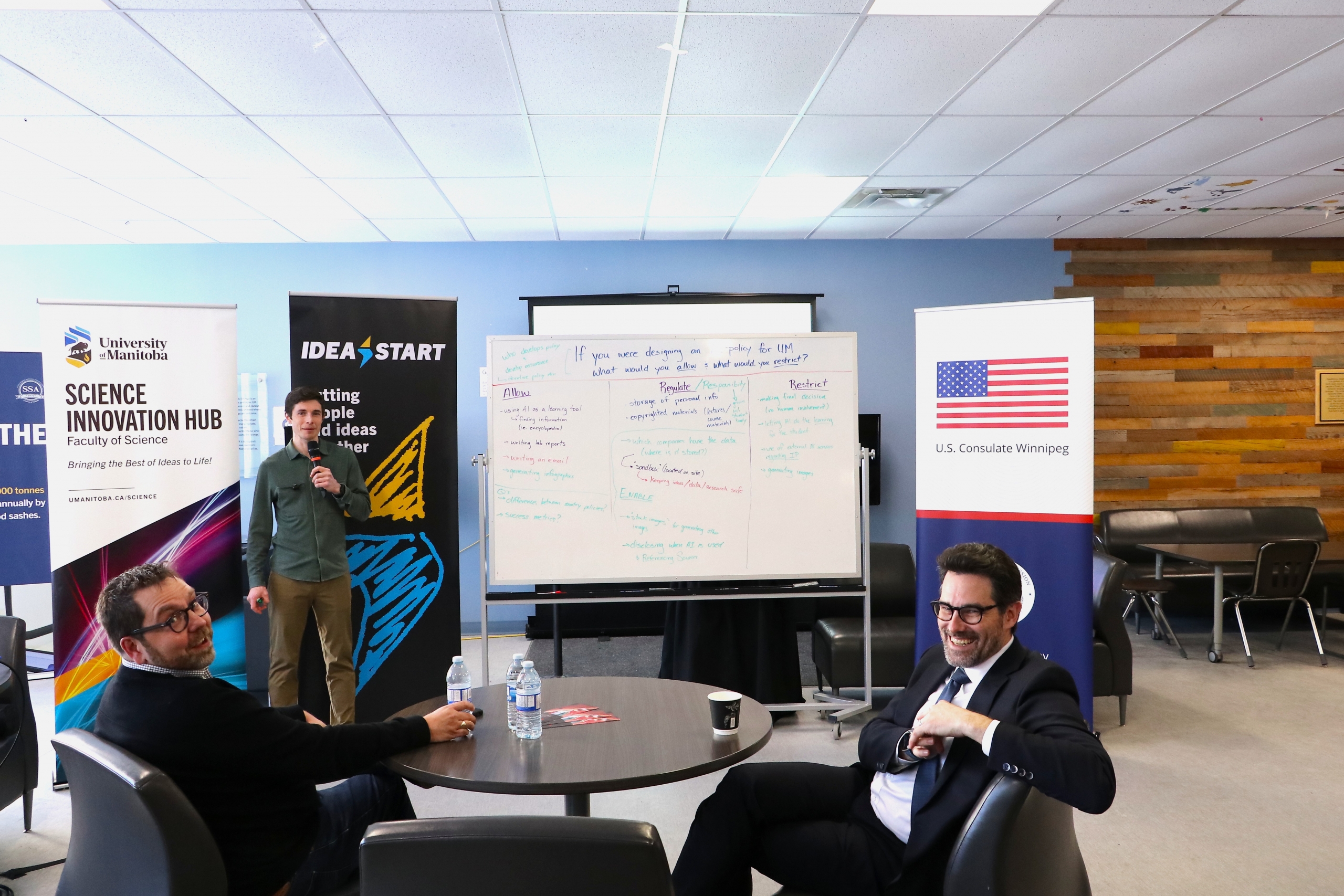 A presenter shares AI policy findings with participants, with University of Manitoba, IDEA START, and U.S. Consulate Winnipeg banners displayed.