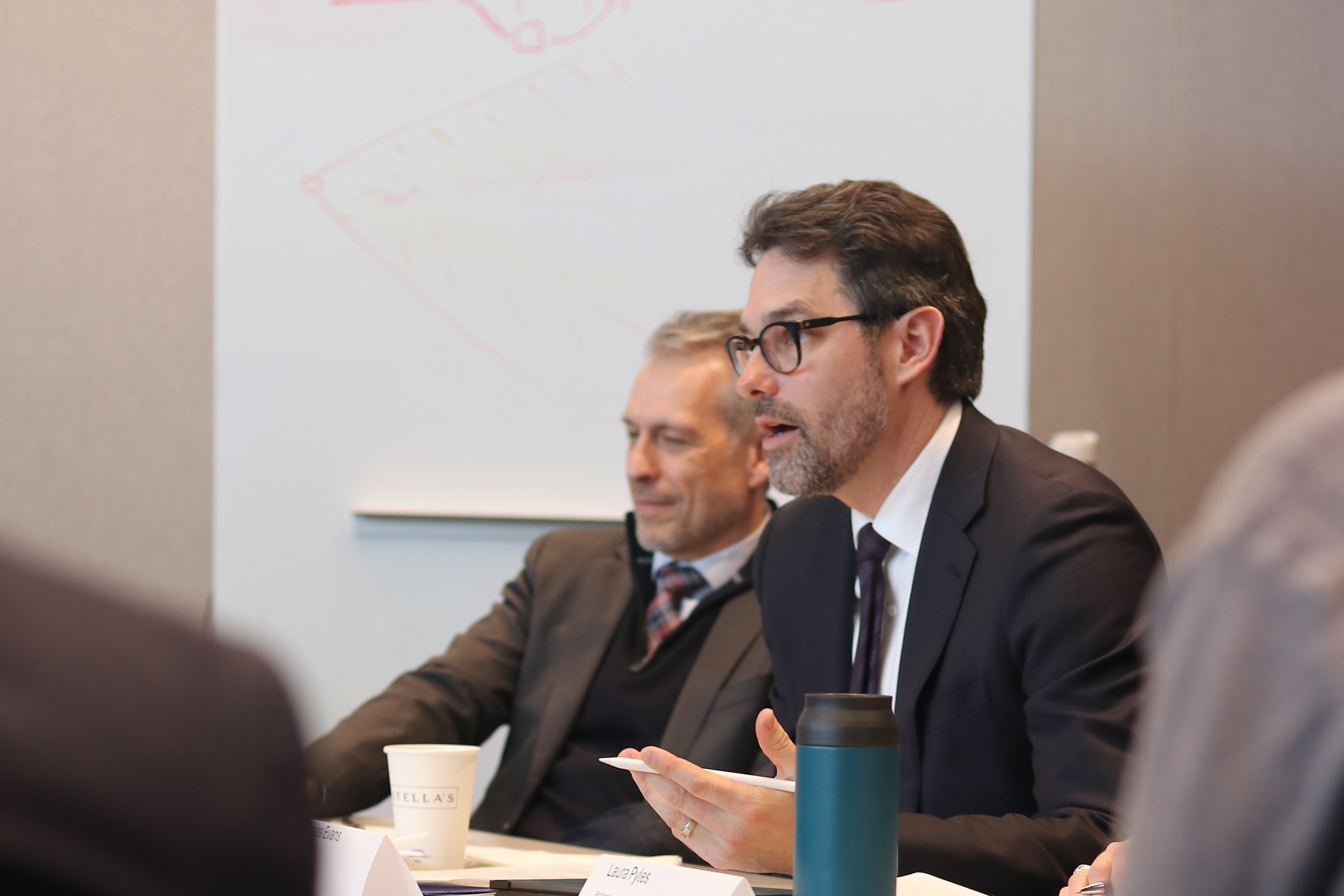 A man in a dark suit gestures while speaking at a roundtable discussion, with a name placard, papers, and coffee visible on the table in front of him.