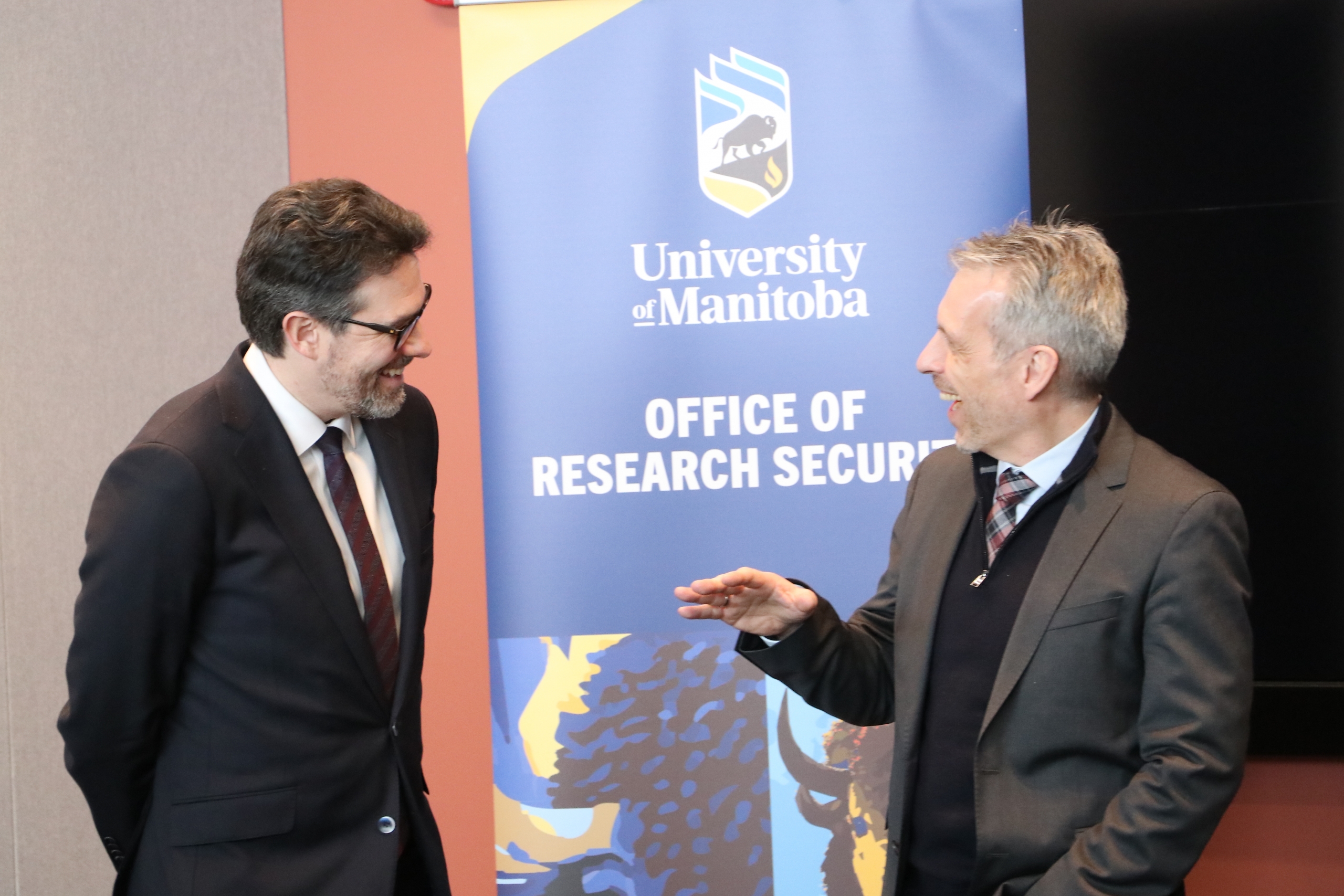Alt text: Two men in suits share a conversation and laugh in front of a University of Manitoba Office of Research Security banner.
