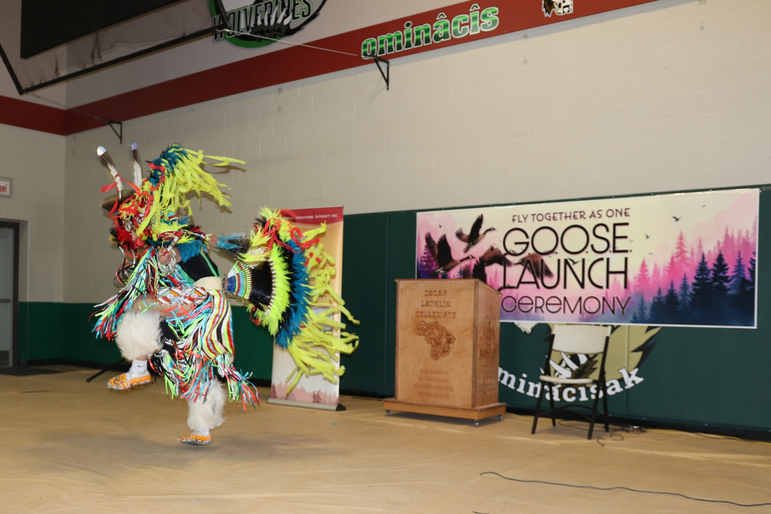 An Indigenous cultural dancer performs in a school gymnasium in front of a sign that reads "GOOSE launch."