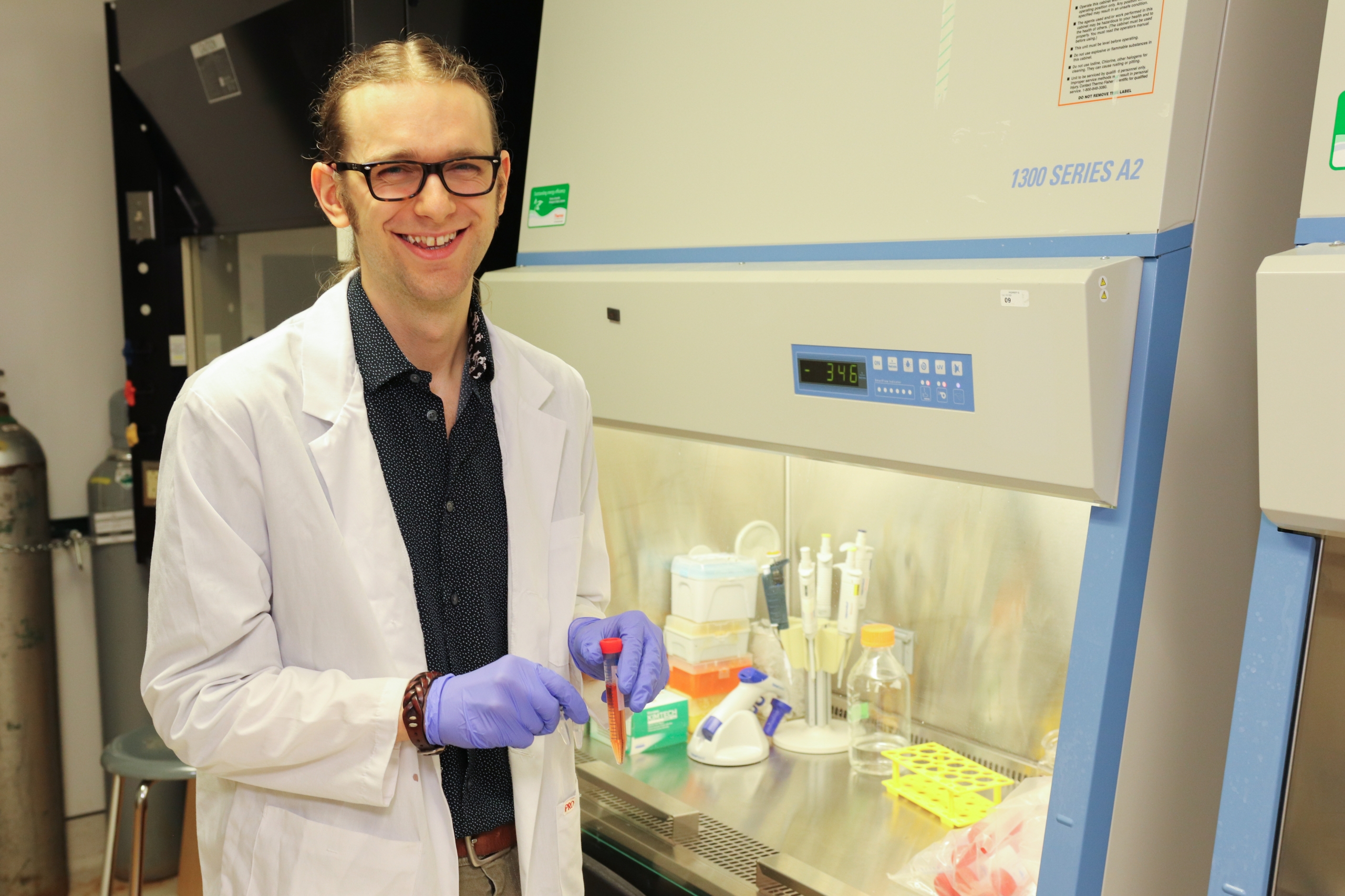 Eli Mondor holds a test tube and smiles at the camera, with scientific equipment visible in the background.