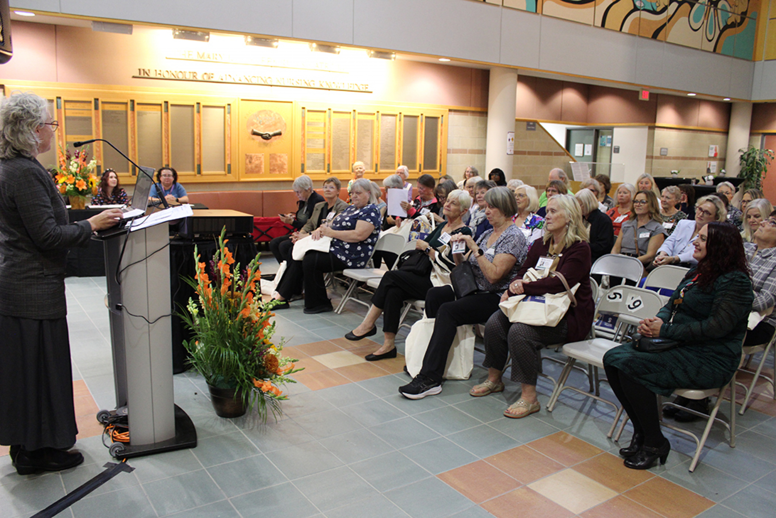 A crowd of people gathered in the College of Nursing atrium, as dean Dr. Kellie Thiessen speaks from a podium.
