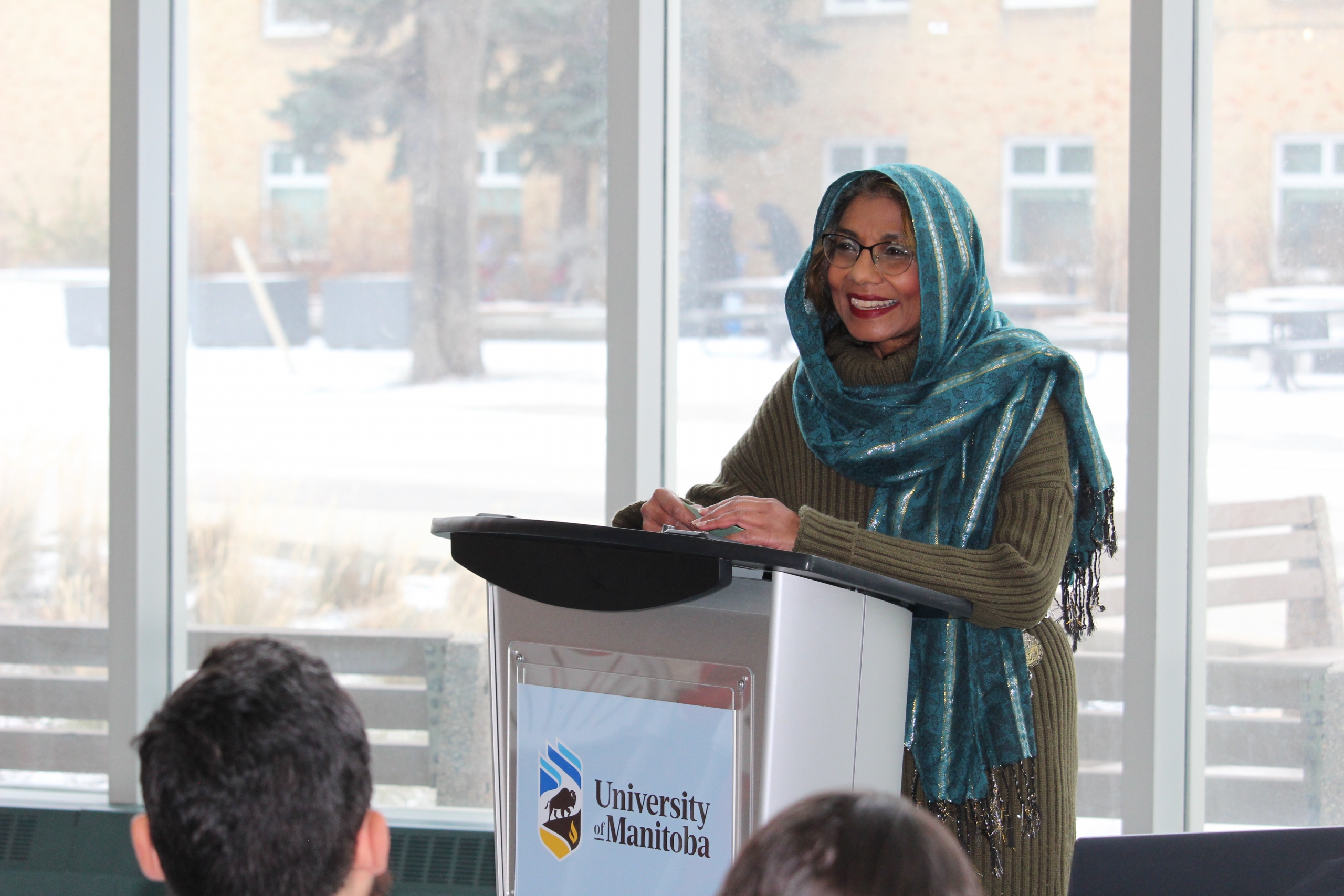 Natasha Ali, UM Muslim spiritual care provider speaks at a podium in front of a large window at Bannatyne campus.
