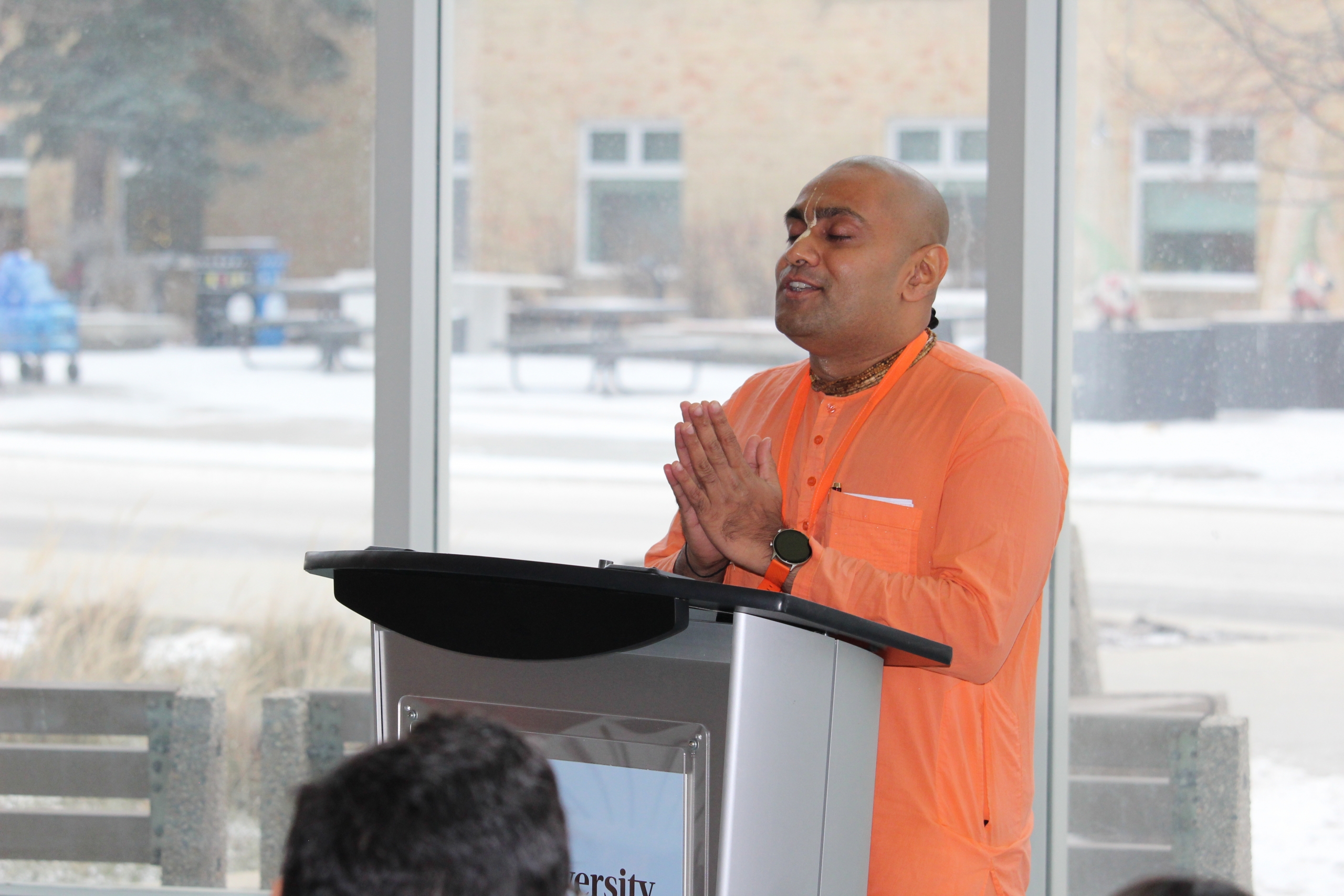 Sanatana Dharma Das stands at a podium at Bannatyne campus, with hands together in prayer.