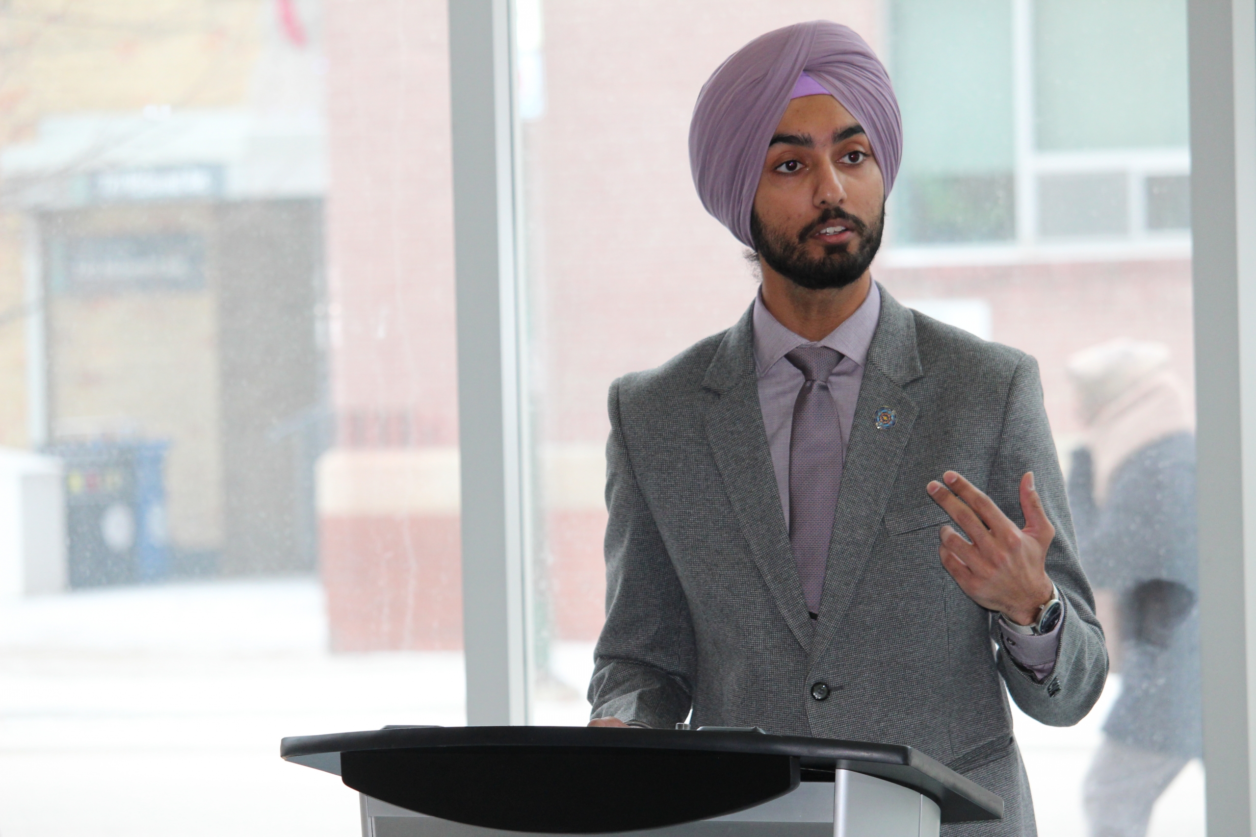 Prabhnoor Singh speaks at a podium in front of a large window at Bannatyne campus.