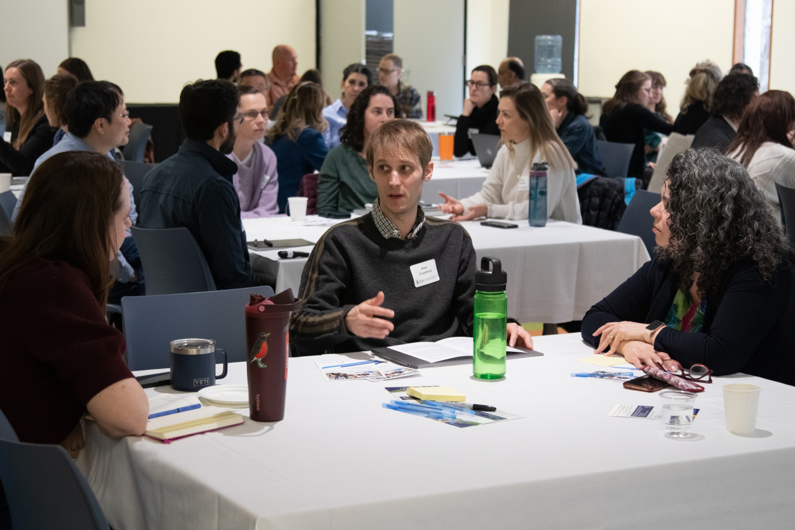 The audience is shown sitting in clusters at their tables, while one group is involved in a discussion.