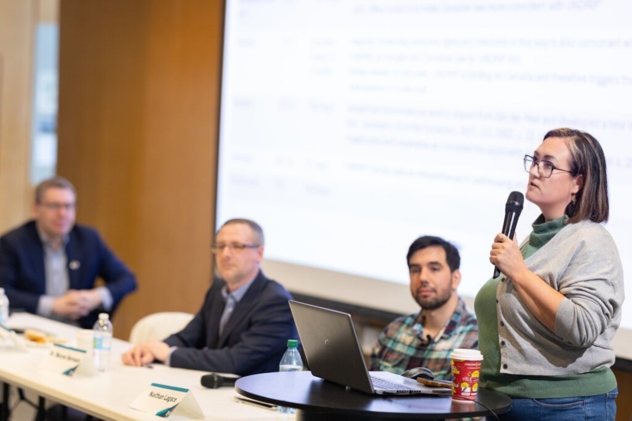 Woman speaking into a microphone at a podium while three panelists sit at a table during an indoor presentation.