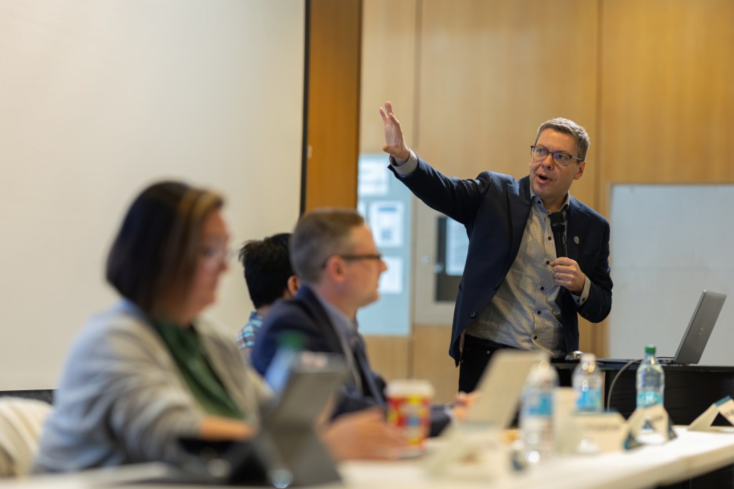 Man holding a microphone gestures toward the screen while panelists sit at a table during an indoor discussion.