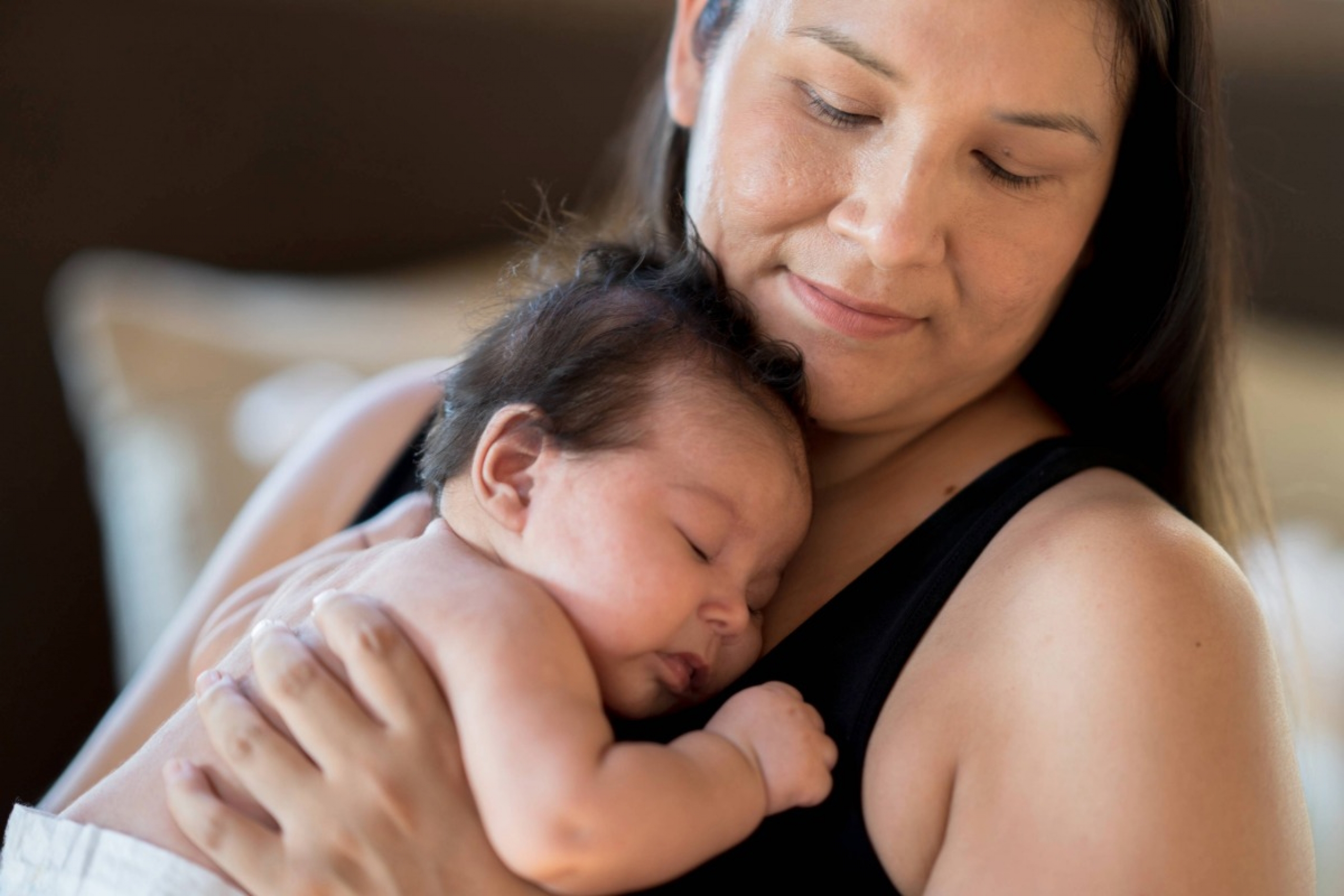 An Indigenous mother sits with her newborn daughter on her chest. The mother is dressed in a black tank top and the baby is wearing just a diaper.