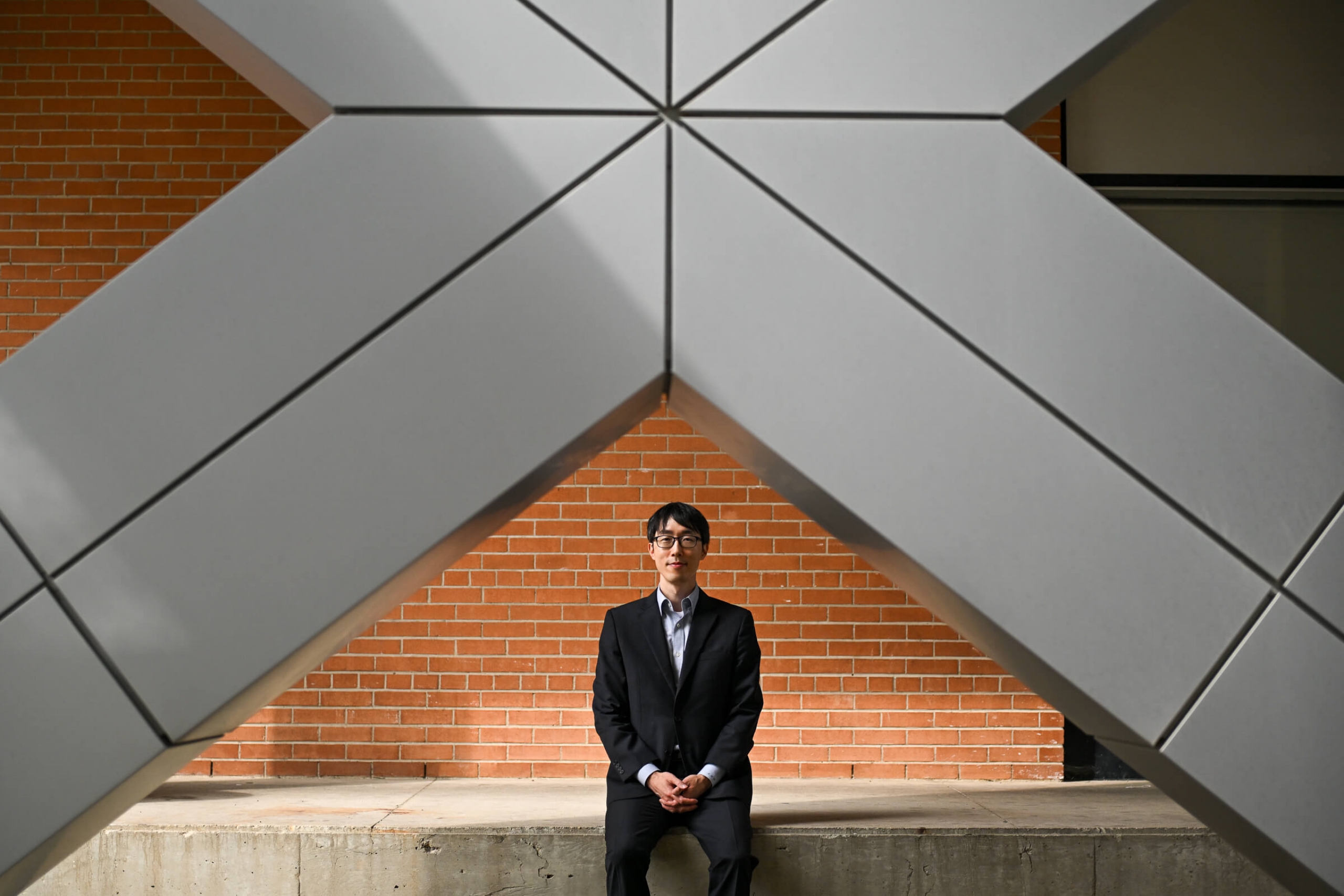 A mean with glasses (Jae Yun Kim) in a suit sitting in front of a building