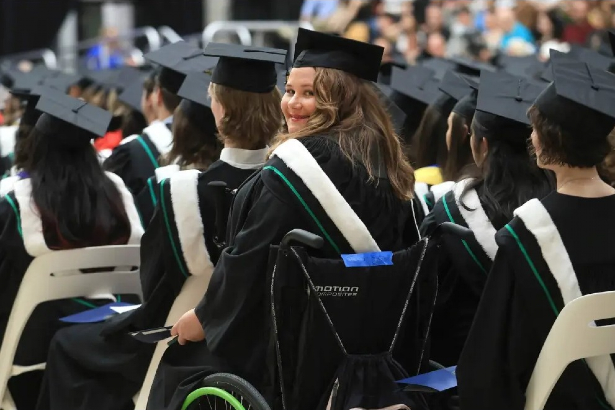 Woman in a wheelchair wearing a graduation cap and gown, looking over her shoulder at the camera.
