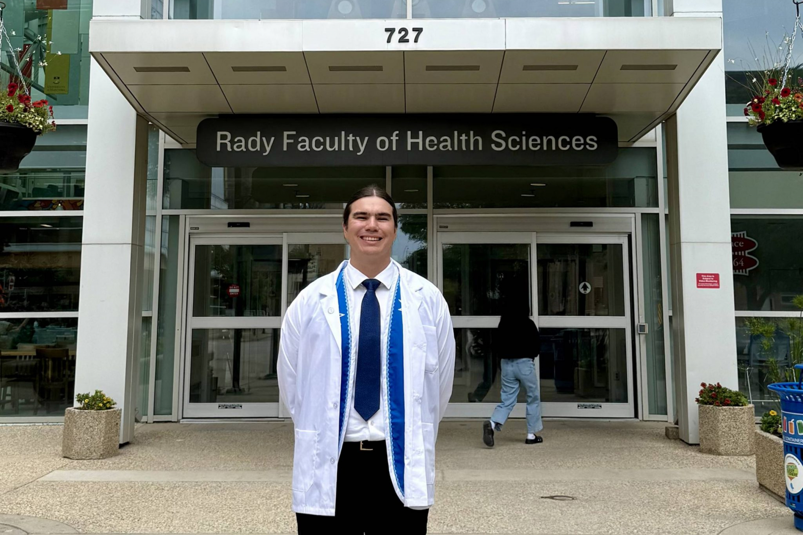 A young smiling man in a white coat in front of a building