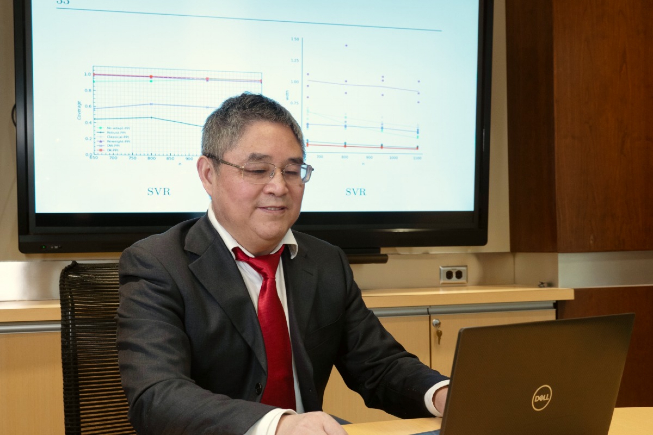 A researcher working on his computer, behind is a screen with graphs.
