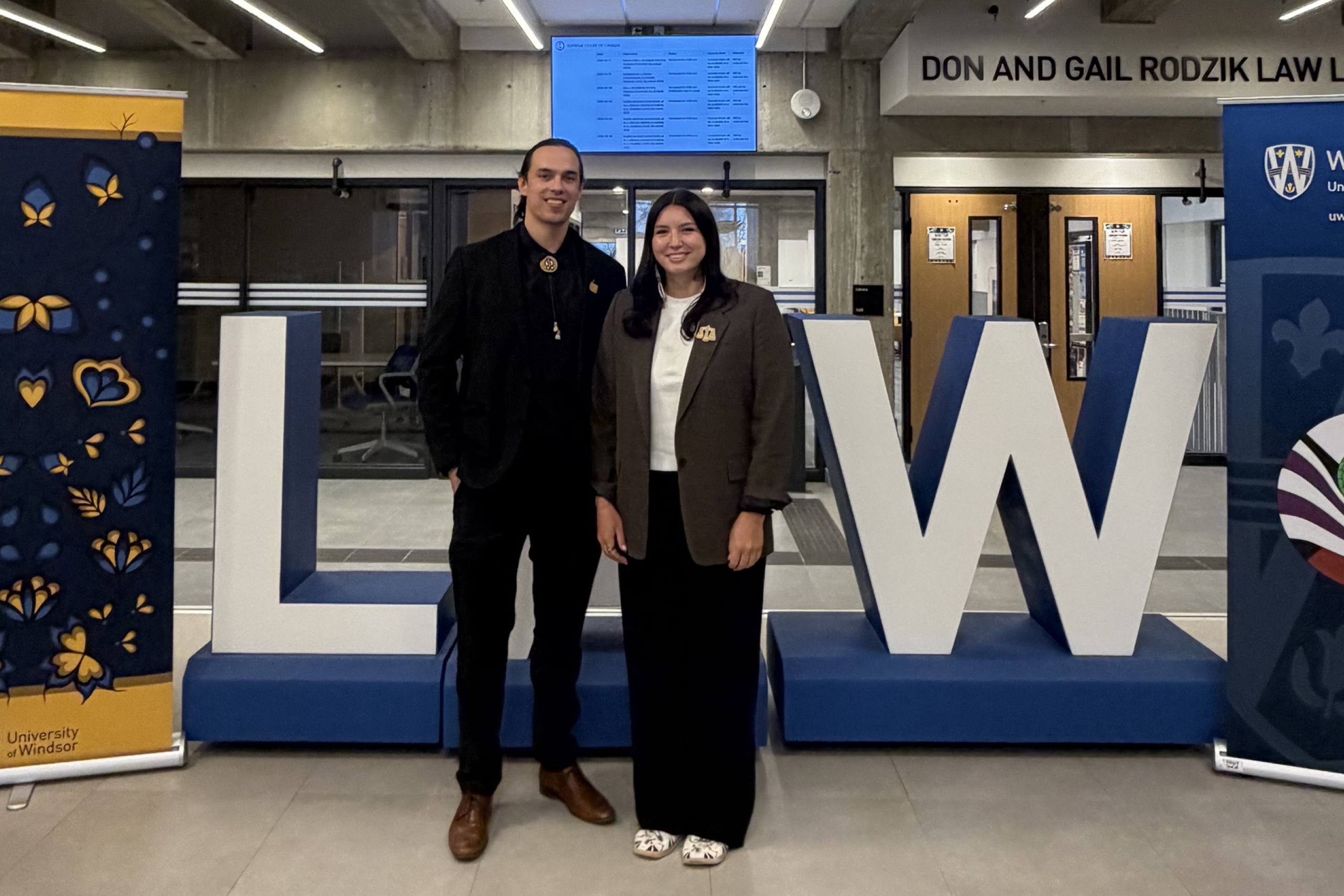 two law students in formal suit jackets and dress pants stand in front of a large sign that says LAW.