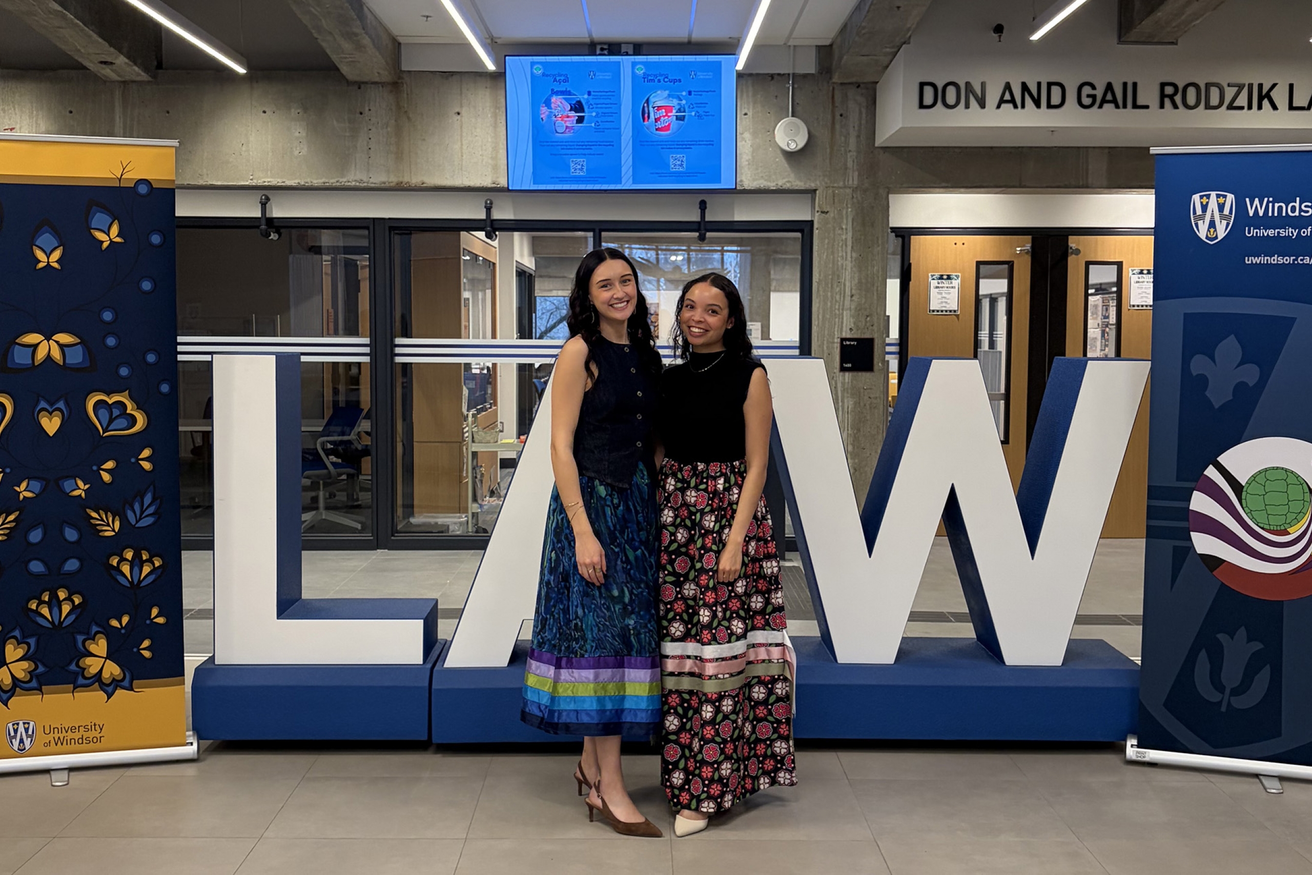 two law students in formal ribbon skirts stand in front of a large sign that says LAW.