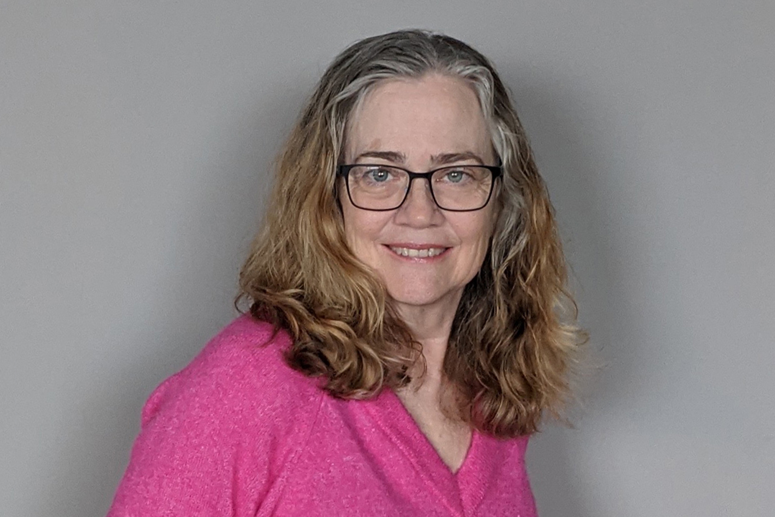 Karen Busby smiles facing the camera wearing a pink sweater. Her hair is shoulder-length blonde with grey at the temples.