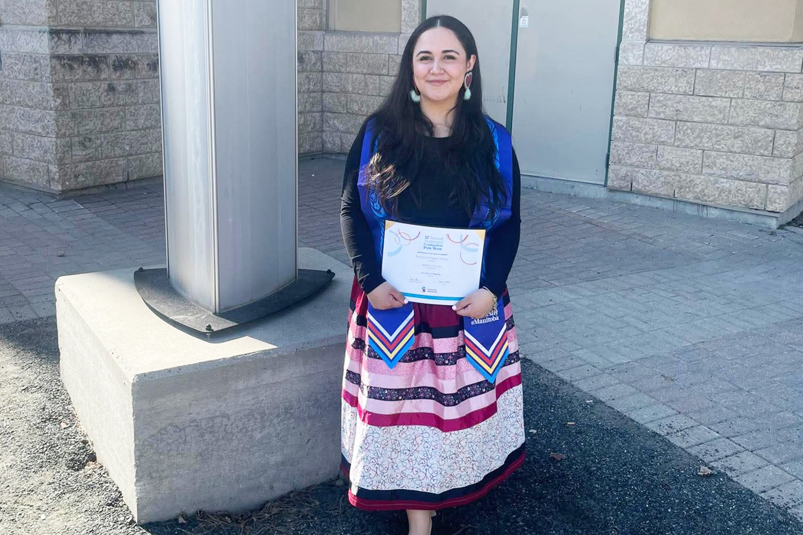 Kaydence Morgado Thomas wearing a ribbon skirt and holding a certificate, while standing outside.