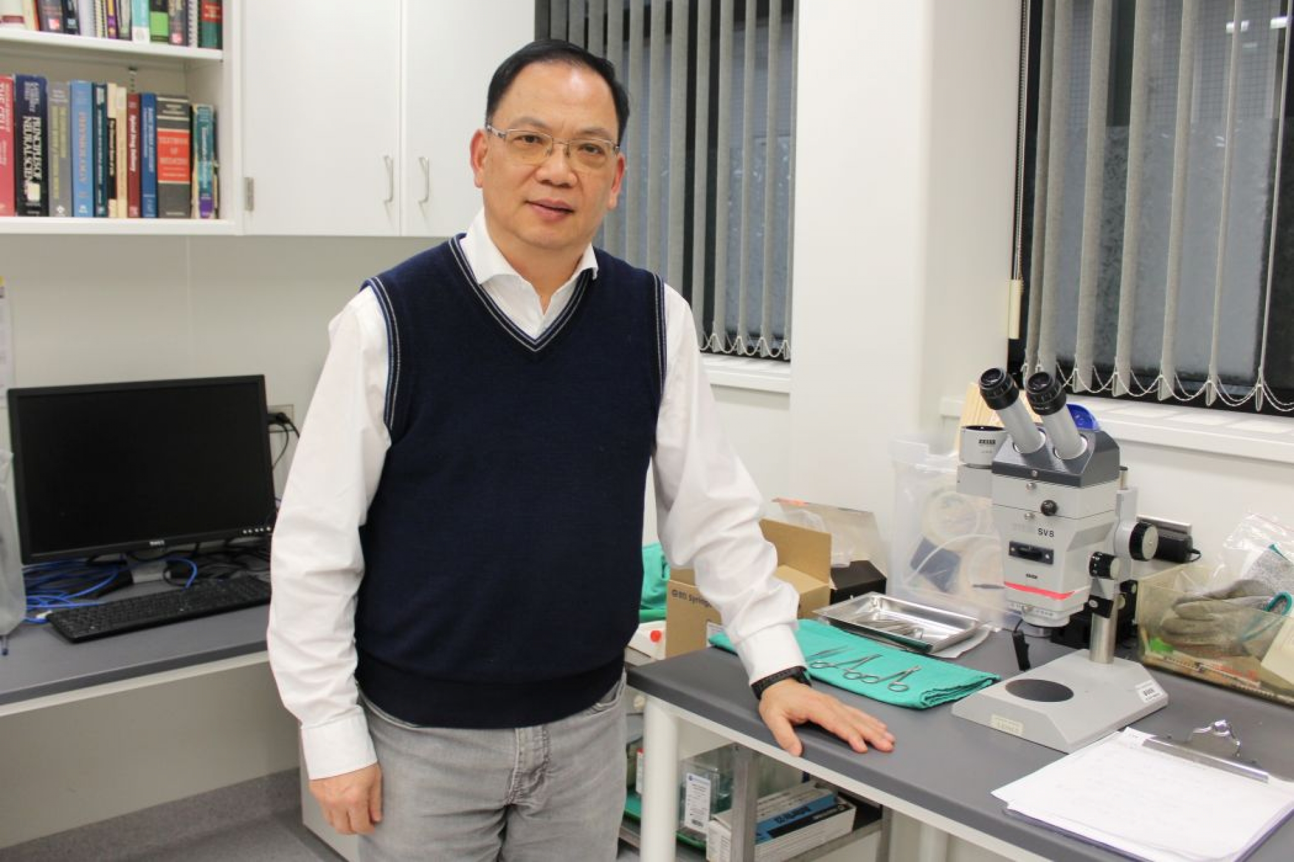Dr. Jiming Kong stands in his lab next to a microscope. A computer and books are in the background.