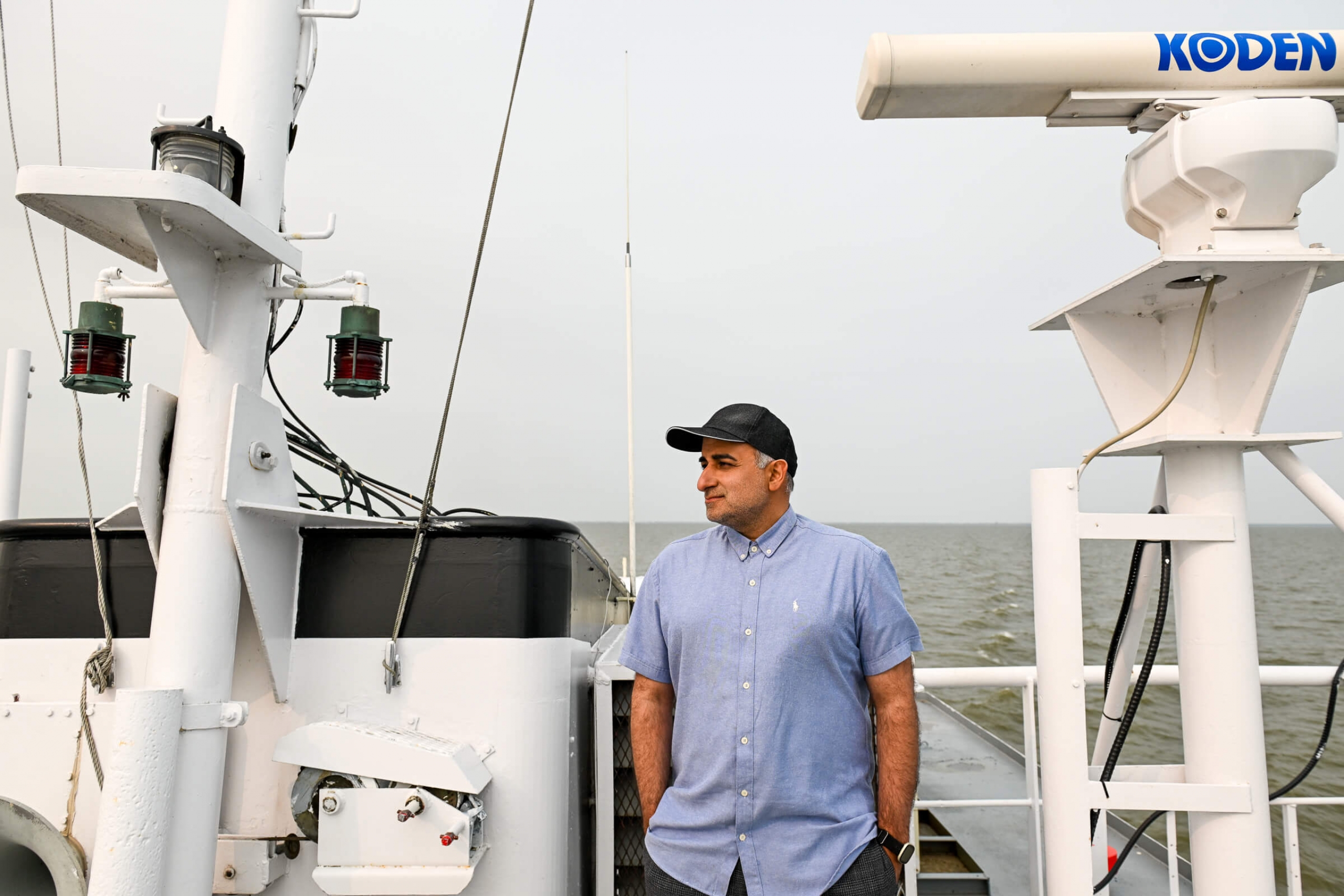 A man (Masoud Goharrokhi) standing on the deck of a ship wearing a black ball cap